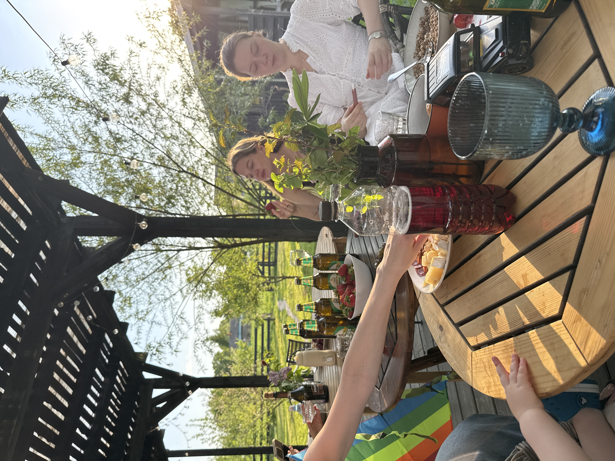 The image shows a group of people gathered around a wooden table, seemingly at an outdoor picnic or gathering in Prudki, Russia. Two women are seated at the table, one wearing a white dress and the other with light-colored hair; both appear to be eating or interacting with items on the table. Various items, including bottles of liquids (possibly wine or juice), glasses, a bowl of strawberries, and a plate with cheese and sausage, are arranged on the table. A child's hand is visible near the edge of the table. The setting appears to be a garden or backyard, with trees and open space in the background. A pergola or wooden structure with lights is visible on the left side of the image.