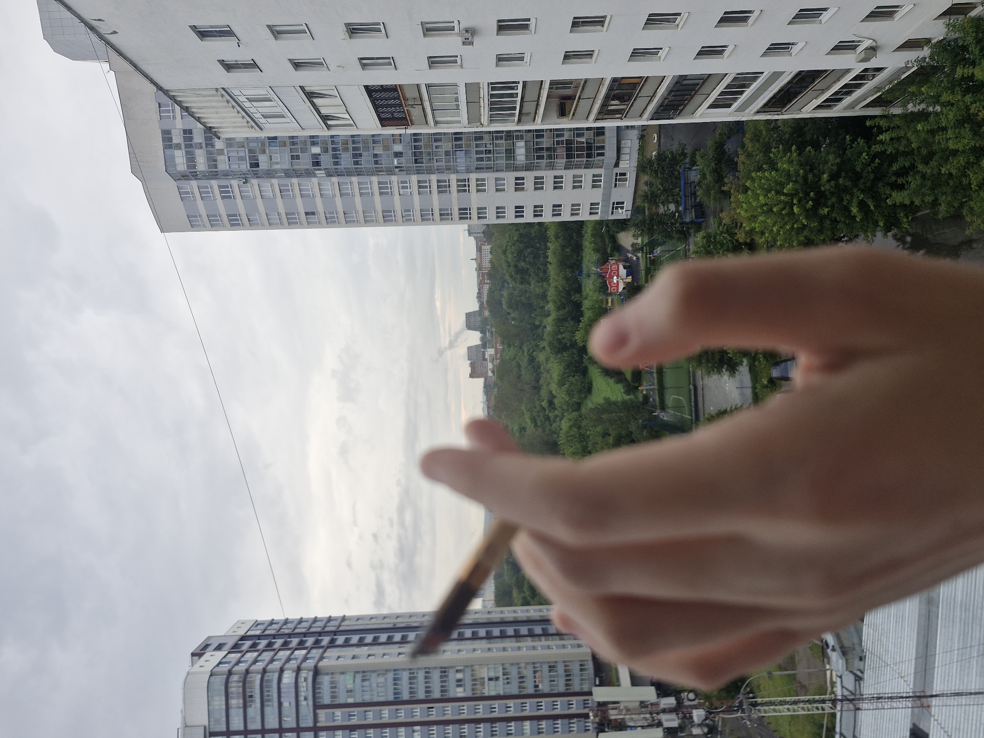 The image shows a hand holding a cigarette in the foreground, with a blurred view of a city skyline in the background. The city appears to be Yekaterinburg, Russia, judging by the architectural style of the buildings and the overall urban environment. The scene is set on an overcast day, with the sky filled with clouds. The buildings are modern high-rises, with some greenery visible between them. There are also some industrial structures and a playground in the distance. The hand is holding the cigarette between the index finger and thumb, with other fingers blurred and out of focus. The cigarette is lit, with a hint of smoke visible.