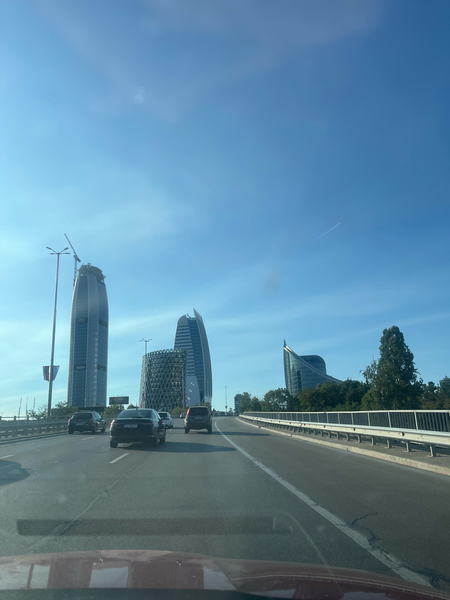 The image shows a view from a vehicle driving on a multi-lane road in Sofia, Bulgaria. In the background, several modern skyscrapers are visible, including a very tall, slender building with a crane at its top, indicating ongoing construction. Another building features a distinctive patterned facade, and a third has a curved, sail-like design. The sky is a clear blue with a faint contrail from an aircraft. Trees line the side of the road, and a white metal guardrail separates the road from an adjacent area. Several cars are on the road ahead, some in the same lane as the viewer and others in adjacent lanes. The overall scene depicts a contemporary urban environment with significant architectural development.