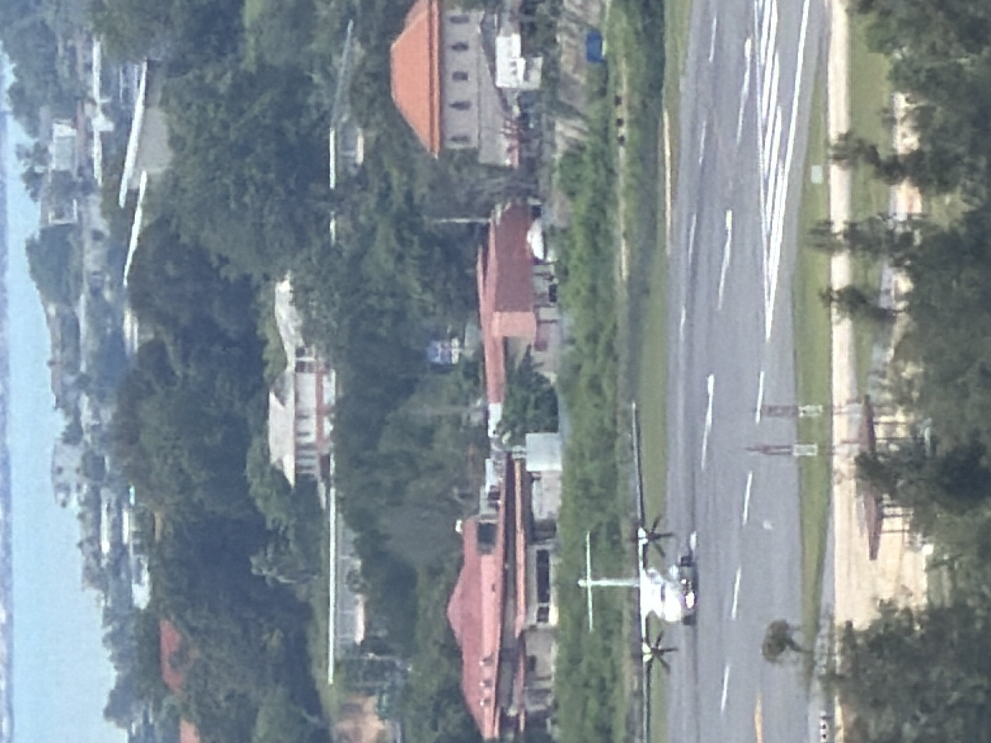 The image depicts an aerial view of a tropical airport with a turboprop airplane on the tarmac in the foreground. The airplane is white with visible propellers on its wings. The tarmac features clear runway markings. In the background, several buildings with red-tiled roofs are scattered among lush green hills. The overall scene suggests a sunny day with clear skies, typical of a coastal or island setting. The presence of tropical vegetation indicates a warm climate. Based on the context provided, this airport is located in Ban Bang Rak, Thailand.