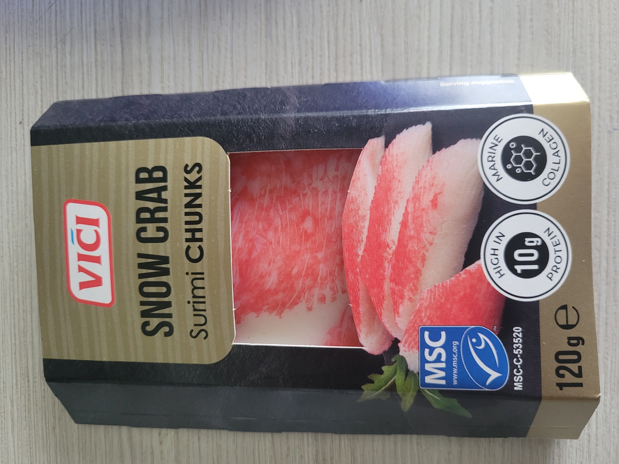The image shows a close-up, overhead view of a package of "Vici Snow Crab Surimi Chunks" placed on a light-colored, textured surface that appears to be wood. The packaging is predominantly black and gold, with a clear window revealing the pink and white surimi. Prominently displayed on the packaging are the brand logo "VICI", the product name "SNOW CRAB SURIMI CHUNKS", an MSC (Marine Stewardship Council) certification logo, a nutritional claim indicating "HIGH IN 10g PROTEIN", a marine collagen icon, and the net weight "120g e". The packaging is slightly angled, with the bottom left corner closer to the viewer. There are no people or specific landmarks visible. The context of Jūrmala, Latvia, is not discernible from the provided image.