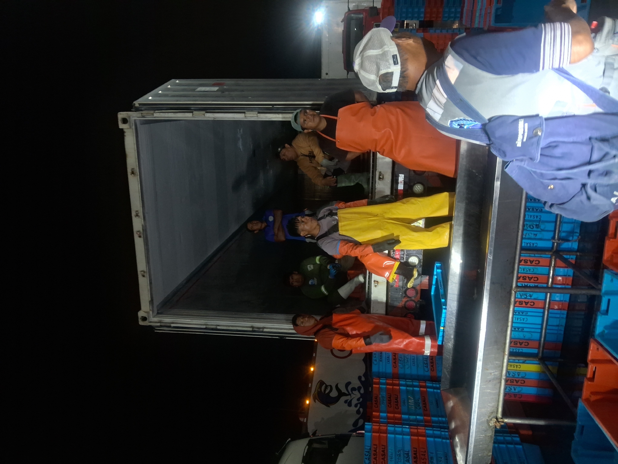 The image depicts a group of people, likely fishermen, standing by an open shipping container at night, in what appears to be a port or market setting in Chimbote, Peru. The individuals are wearing various types of protective gear, including bright orange and yellow overalls, aprons, and gloves, suggesting they are involved in handling fish or seafood. Stacks of blue and red plastic crates are visible in the foreground and background, further indicating a commercial fishing operation. The lighting suggests it is nighttime, with artificial lights illuminating the scene. One man in the foreground is wearing a grey vest and a blue backpack. The shipping container is partially loaded or unloaded, with people inside and around it. The overall atmosphere is one of active, late-night work in a maritime industrial environment.