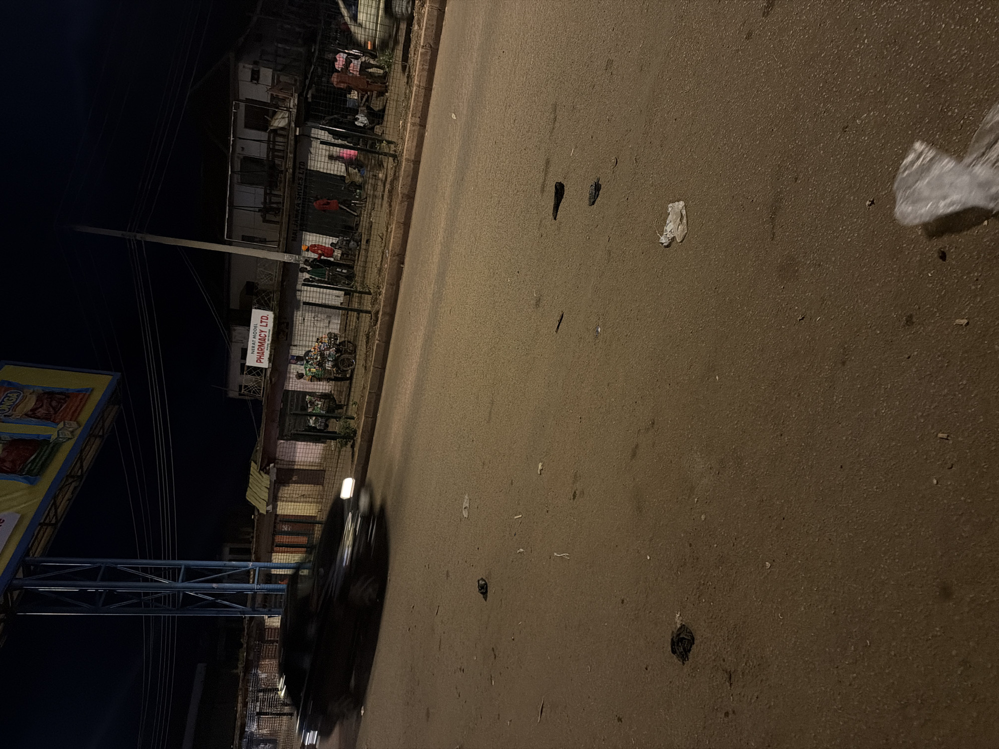 A dark-colored sedan is captured in motion on a paved road at night in Bukuru, Nigeria. The vehicle's headlights are illuminated, indicating it is moving. The road surface is rough and appears to be in need of repair, with debris scattered across it, including plastic bags, crumpled paper, and dark organic matter. In the background, a row of low-rise buildings lines the opposite side of the road. Signs indicating "NERAT MODEL PHARMACY LTD." and "MAI JAMA'A SHOES LTD." are visible on these structures, suggesting a commercial area. Several people are gathered in front of the buildings, some standing and others appearing to be sitting. A motorcycle with a cargo rack is parked near the pharmacy. Overhead, electrical wires are strung across the dark sky, and a large advertising billboard, partially illuminated, is visible. The overall atmosphere is that of a busy, urban environment at night, characterized by vehicular traffic and pedestrian activity.