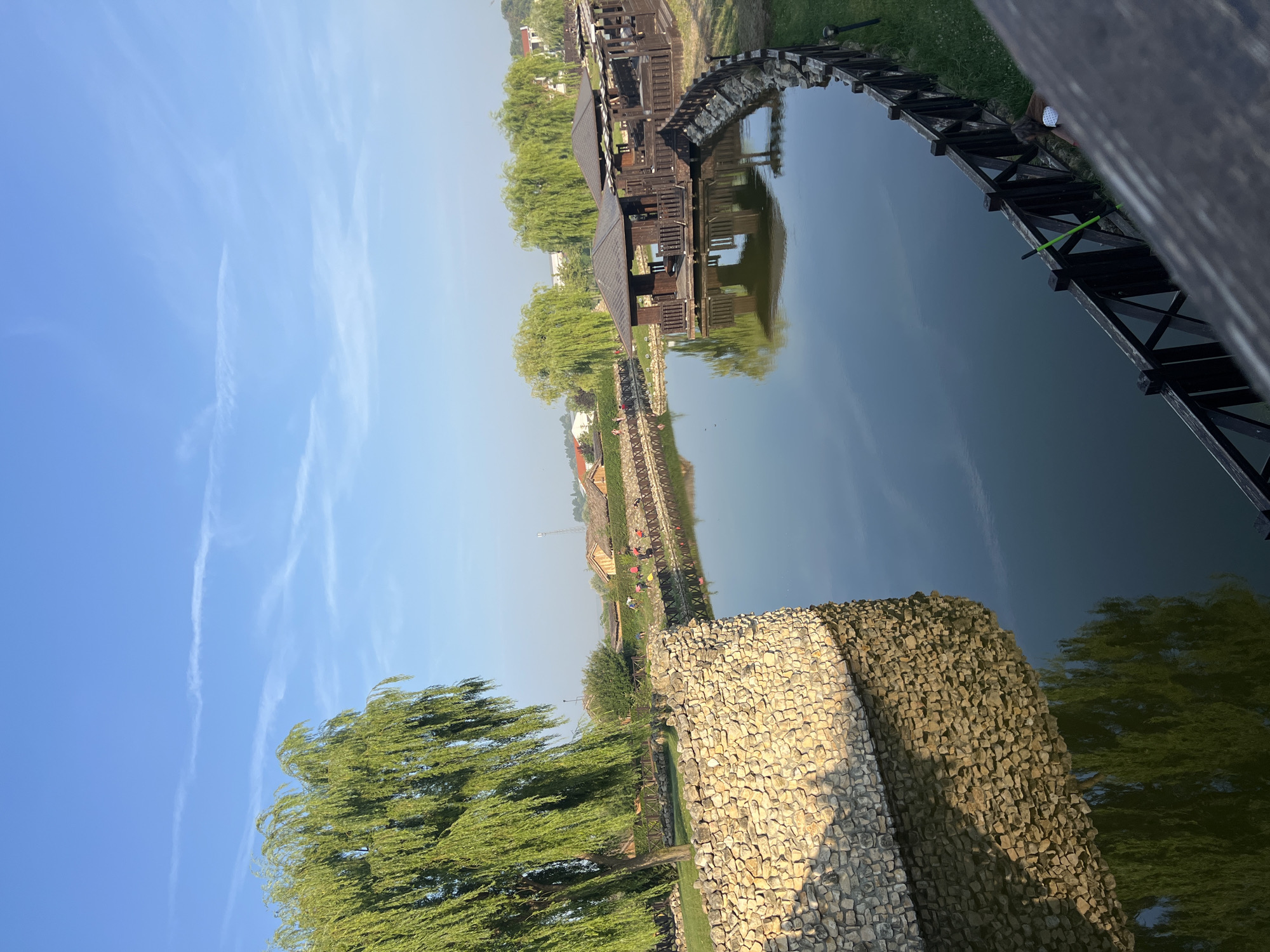 A picturesque view of a serene lake is captured from an elevated perspective, showcasing a tranquil environment. The lake's surface, smooth and reflective, mirrors the clear blue sky adorned with wispy white clouds. Along the edge of the lake, a charming stone wall borders a lush green lawn, its texture and form elegantly reflected in the water. Beyond the wall, a majestic weeping willow tree stands tall, its verdant branches cascading downwards, also reflected in the lake.
Further along the lake's perimeter, a series of rustic wooden gazebos are built over the water, offering shaded retreats. A dark wooden fence lines the walkway that encircles the lake, separating the water from the surrounding grassy areas and a few scattered trees. In the background, several buildings with thatched roofs are visible, suggesting a village or resort setting. A few people can be seen walking along the path and near the buildings. A person is crouching by the fence on the right side of the image, possibly tending to something. The overall scene evokes a sense of peace and natural beauty, a perfect spot for relaxation and leisure. The location is in the city of Neofit Rilski, Bulgaria.