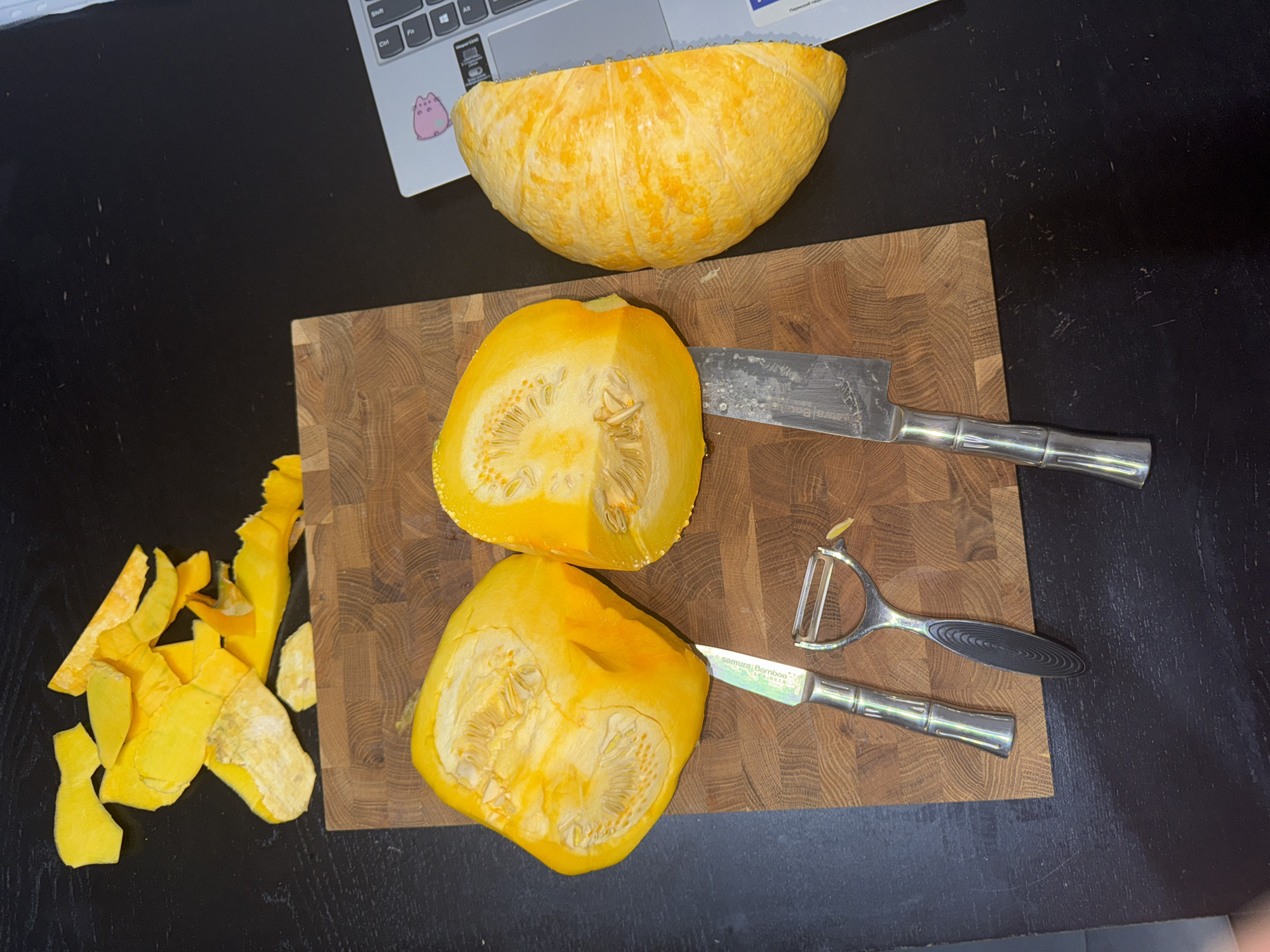 A high-angle shot shows a kitchen scene with a dark, wooden countertop. In the center, a butcher block cutting board is laid out. On the board, two halves of a bright yellow squash are visible, revealing their seeds and fibrous interiors. A vegetable peeler and two knives are also placed on the cutting board. To the left of the board, yellow squash peelings are scattered. To the right of the cutting board, a portion of a silver laptop with a pink sticker on it is visible. The scene is well-lit and appears to be indoors.