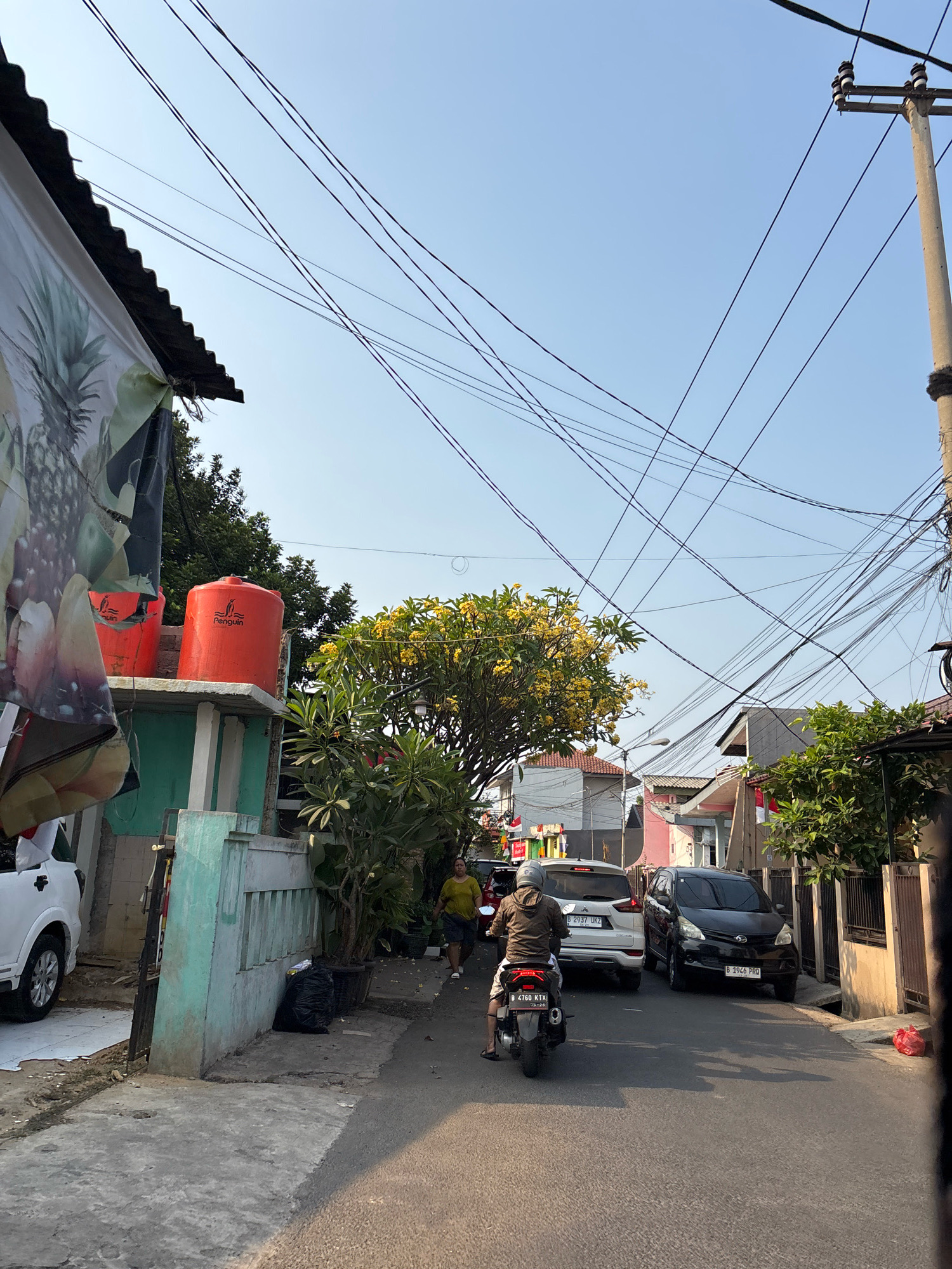 The image depicts a street scene in Bekasi, Indonesia, under a clear sky. A narrow asphalt road runs through a residential area lined with houses and buildings. Overhead, a complex network of electrical wires crisscrosses the sky, supported by a concrete utility pole on the right.
In the foreground, a person on a motorcycle, wearing a helmet and a brown jacket, is moving away from the camera. The motorcycle has a license plate that reads "B 4760 KTX". To the left of the road, a white SUV is parked. Further down the street, a white car and a black car are also visible. The black car has a license plate "B 1946 PRO".
On the left side of the street, a building has a large banner featuring an image of pineapples and other fruits. Two orange water tanks are placed on top of this building. Green and light blue painted walls are also visible on this side. Adjacent to the building, a small tree with yellow flowers is in bloom.
On the right side of the street, houses with fences and trees are seen. A red plastic bag lies on the side of the road. The overall impression is of a typical urban street in Indonesia, with evidence of daily life and infrastructure.