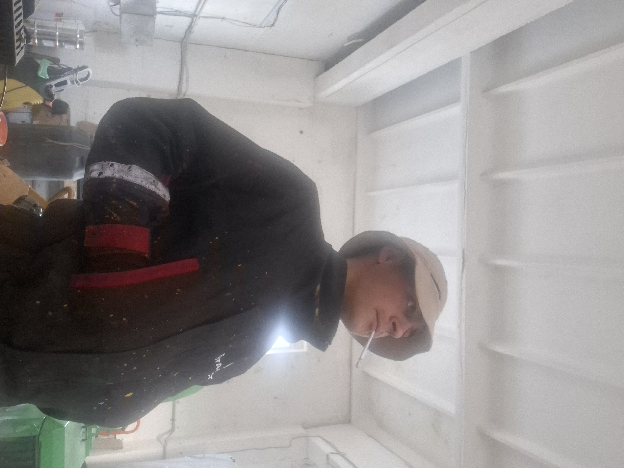 A man wearing a hat and a dark jacket with red accents and white markings is standing in a white room with horizontal moldings. He has a cigarette in his mouth. A green object and tools are visible in the background. The location is in the city of Unknown, Unknown.