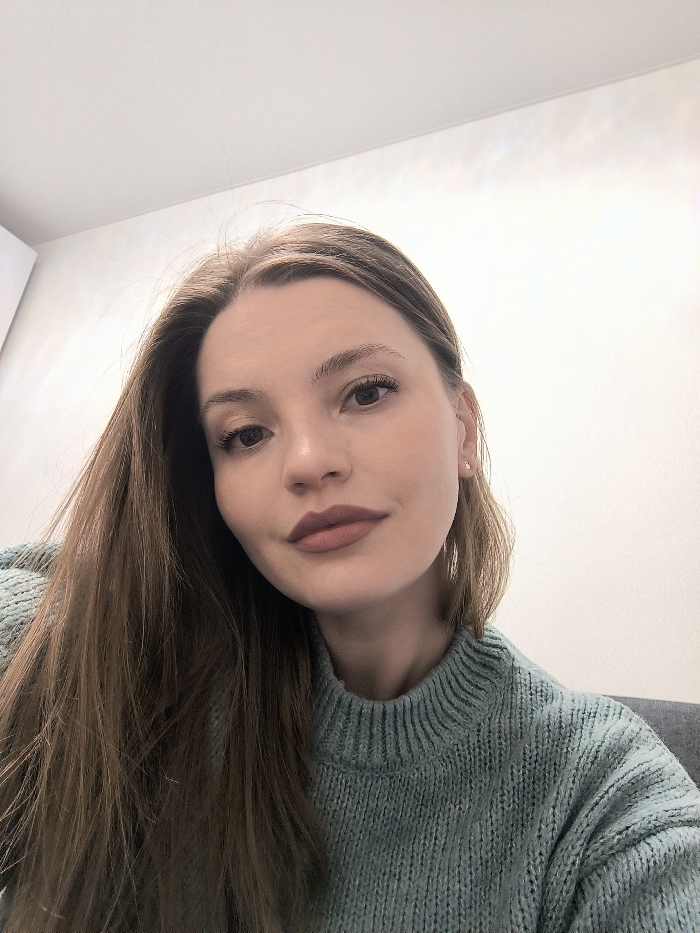 A woman with long, light brown hair is wearing a teal, cable-knit sweater. She has fair skin, light eyebrows, and brown eyes. Her lips are a mauve shade. The background is a plain white wall with some shadows in the upper right corner. She is looking directly at the camera. The framing is a medium shot, focusing on her face and upper body. A hint of a gray or dark couch is visible in the lower right corner.