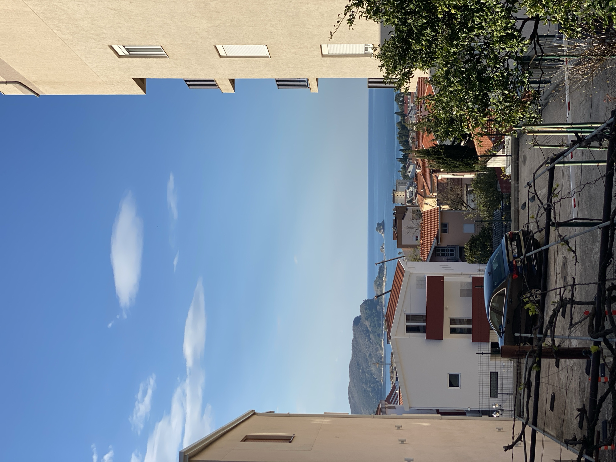 The image shows a street scene in Budva, Montenegro. Buildings line both sides of the frame, creating a narrow perspective toward the sea. The sky is bright blue with scattered white clouds. Closer to the viewer, the scene is slightly obscured by greenery, including a tree and a vine growing on a trellis. On the right side, a dark-colored car is parked next to a white house with red trim. The buildings in the distance are a mix of residential and commercial structures, with red-tiled roofs. The sea stretches out to the horizon, with a small island or rock formation visible in the distance. The light suggests it is daytime, with clear weather.