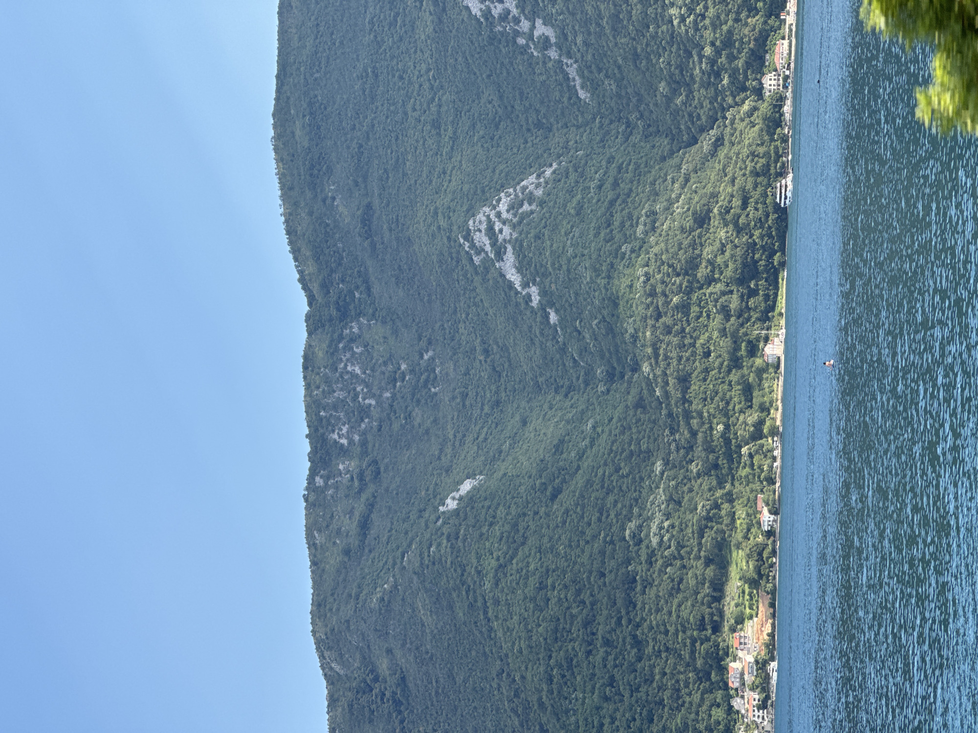 A wide shot captures a sunlit, clear blue sky above a verdant, tree-covered mountain range. The mountains slope down to meet a tranquil body of water, likely the Bay of Kotor, where a few buildings and houses are nestled along the shore. In the foreground, the water is a deep blue with small ripples, and a lone individual can be seen on a paddleboard near the center of the frame. Several boats are visible further out on the water, and some of the buildings appear to be hotels or residences. The scene suggests a peaceful summer day in Lipci, Montenegro.