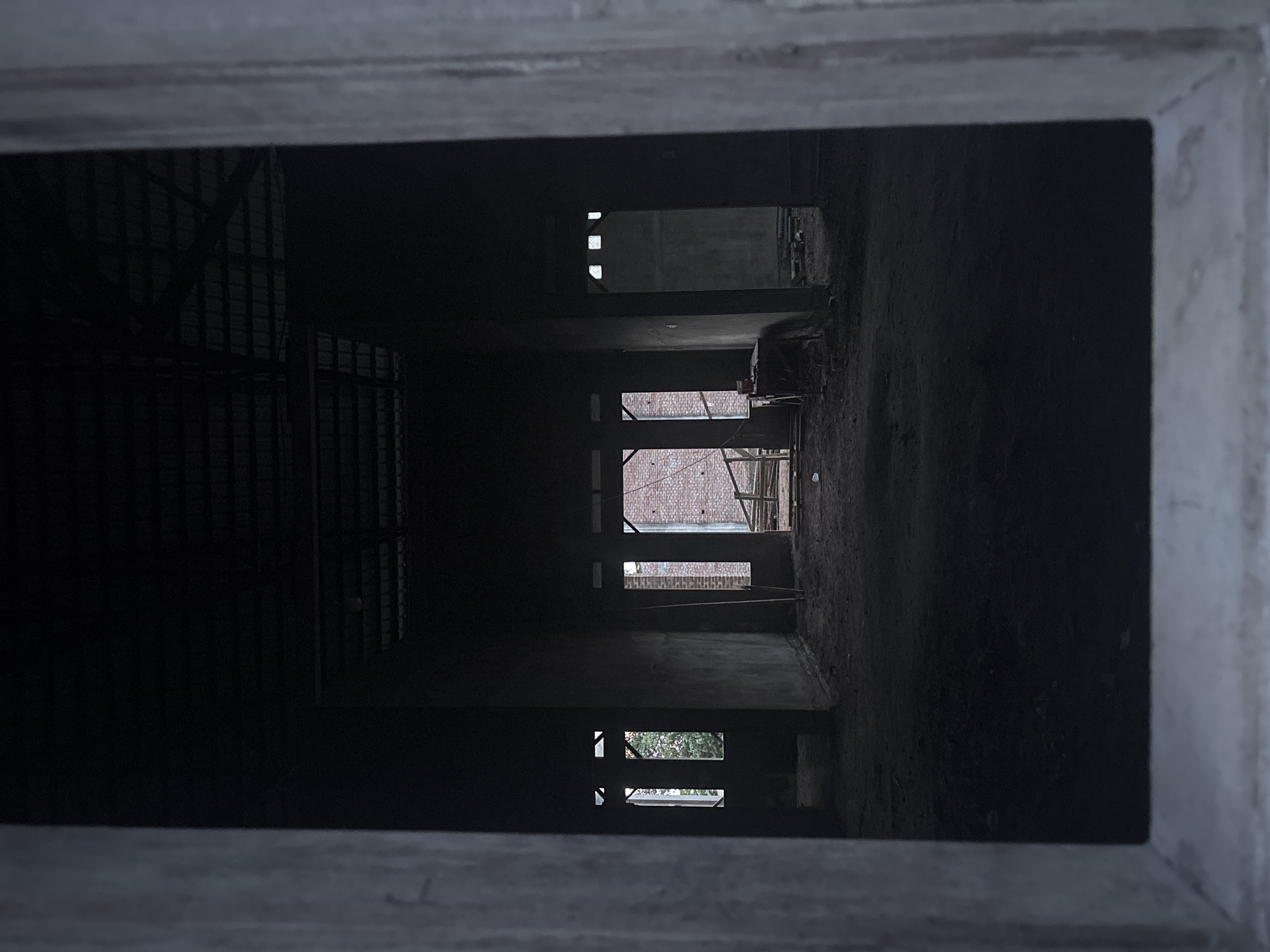 The image shows a perspective shot through a concrete frame into the interior of a building under construction. The interior space appears to have multiple floors, with window openings visible at varying heights. The structure is primarily concrete, with some evidence of brickwork or other building materials visible through the windows. There are no people visible in the image. The environment seems dimly lit and dusty, indicative of a construction site. The location is Cutneuheun, Indonesia.