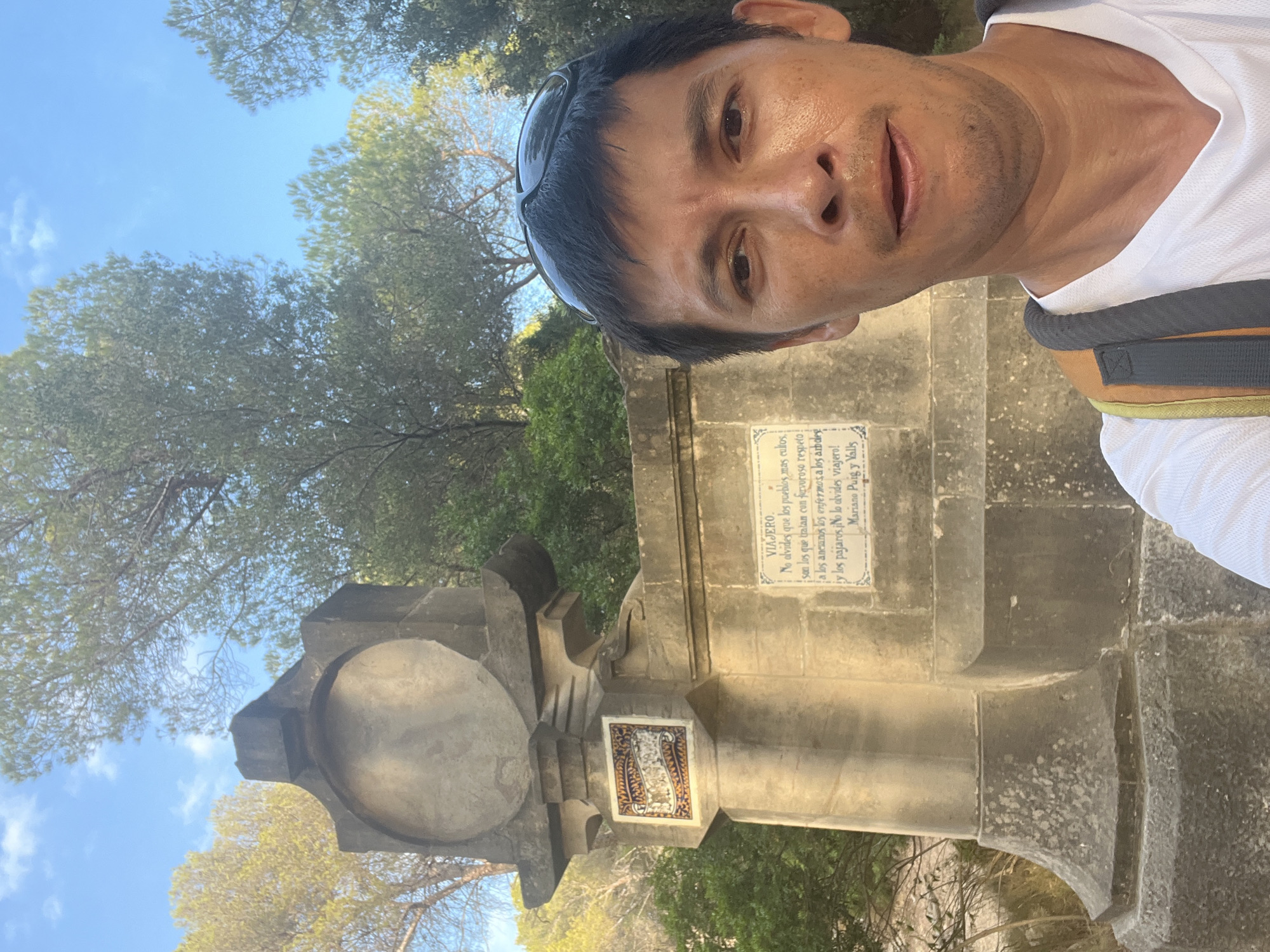 In Tarragona, Spain, a man with dark hair and sunglasses perched on his head takes a selfie. He is wearing a white t-shirt and a backpack with orange and gray straps. Behind him, a stone monument features a circular indentation at the top and decorative tiles on its lower section. A plaque on the monument contains text in Spanish, reading: "VIAJERO. No olvides que los pueblos mas curiosos son los que tratan con fervoroso respeto a los ancianos, los enfermos, a los que viven y los pájaros. No lo olvides viajero!" which translates to "TRAVELER. Do not forget that the most curious peoples are those who treat the elderly, the sick, those who live, and the birds with fervent respect. Do not forget it, traveler!". The monument is surrounded by lush green trees and foliage, with a clear blue sky overhead.