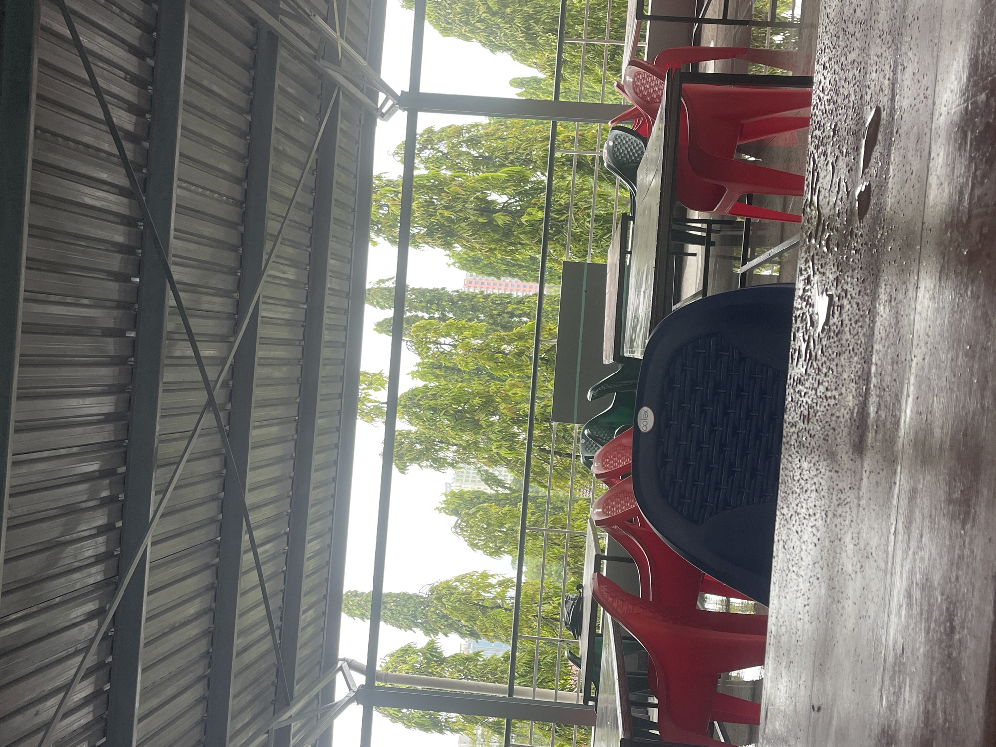 The image depicts a semi-outdoor seating area, characteristic of a cafeteria or restaurant, located in Dar es-Salaam, Tanzania. The perspective is from within the covered space, looking outwards. Overhead and to the left, the underside of a metal roof structure is visible, featuring corrugated sheets supported by a framework of beams.
In the immediate foreground, a wooden table surface is heavily dotted with water droplets, strongly suggesting recent rain or high humidity. Several plastic chairs are arranged around tables in the mid-ground. Most chairs are red, but a black chair with a textured backrest, bearing the faint word "colex," is also visible, along with parts of green chairs.
Beyond the seating area, a verdant landscape of numerous trees stretches into the distance. Through the dense foliage, a tall, light-colored building can be seen. The bright light filtering into the scene indicates it is daytime. No people are present, lending the area a quiet, unoccupied atmosphere, potentially after a rain shower.