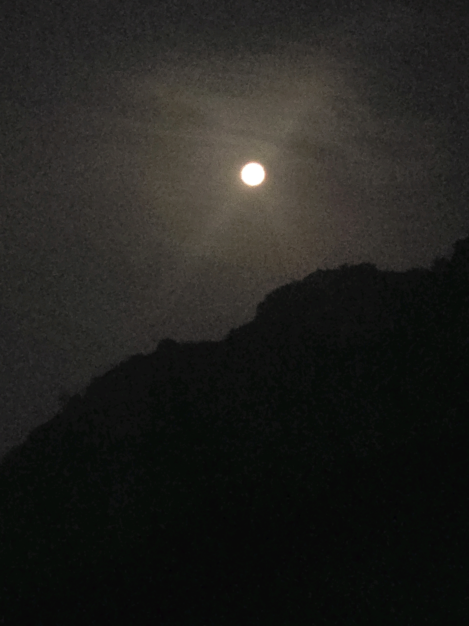 A full moon is visible in the upper central portion of the image, partially obscured by clouds, and casting a soft glow. Below the moon, the dark silhouette of a mountainous or hilly terrain is prominent, with an uneven, jagged skyline. The foreground and much of the sky are dark, with a grainy texture throughout the image, suggesting it was taken at night with low light conditions, possibly with a mobile phone camera. The scene depicts a natural landscape against a dimly lit sky. There are no people or man-made structures visible, nor any indication of its specific location within Podgorica, Montenegro.