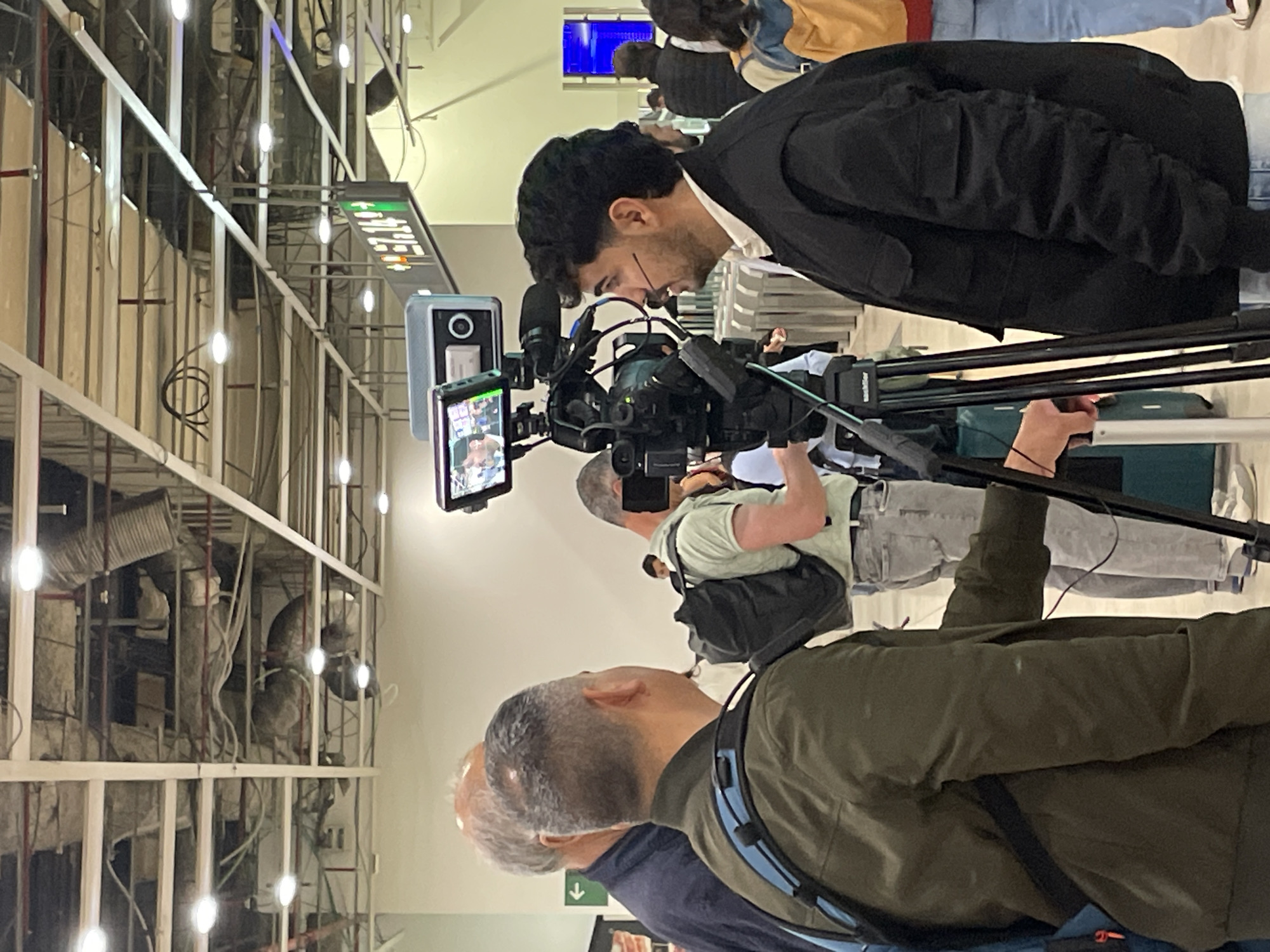 The image depicts a scene indoors, possibly an airport or transportation hub, as suggested by the overhead signage and metal framework visible on the left side of the frame. There are several people present, including a man in a black suit looking into a large camera on a tripod, two other men who appear to be cameramen or assistants, and a few other individuals in the background, partially visible. The camera setup is the prominent subject, with various components like a screen, microphone, and cables. The environment suggests ongoing construction or renovation, given the exposed infrastructure and building materials. The specific location cannot be determined from the image, however, it is indicated that the image comes from the city of Barcelona, Spain.