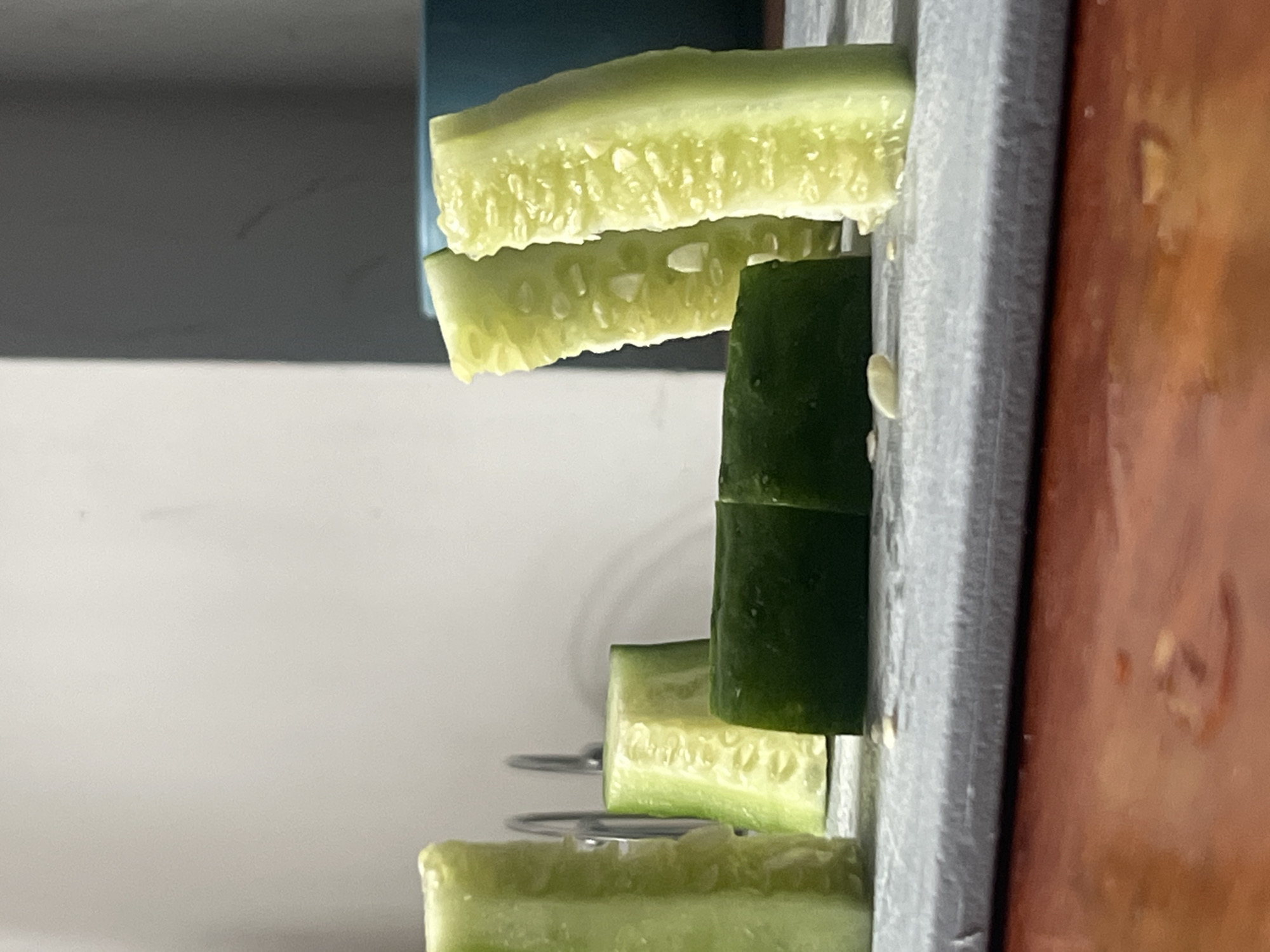 A close-up, indoor shot depicts cucumber slices being processed by a slicer. The cucumber, appearing in various stages of being sliced, presents a contrast between its dark green outer skin and its pale, seed-filled interior. The slicer is a gray, industrial-looking object with wire supports visible beneath some of the cucumber slices. The setting, though not explicitly detailed in the image, is assumed to be located in a kitchen given the presence of food preparation equipment. The image does not show any people or specific interaction, it is a static representation of cucumbers being sliced. There is no information to confirm that this image was taken in Kommunarka, Russia.