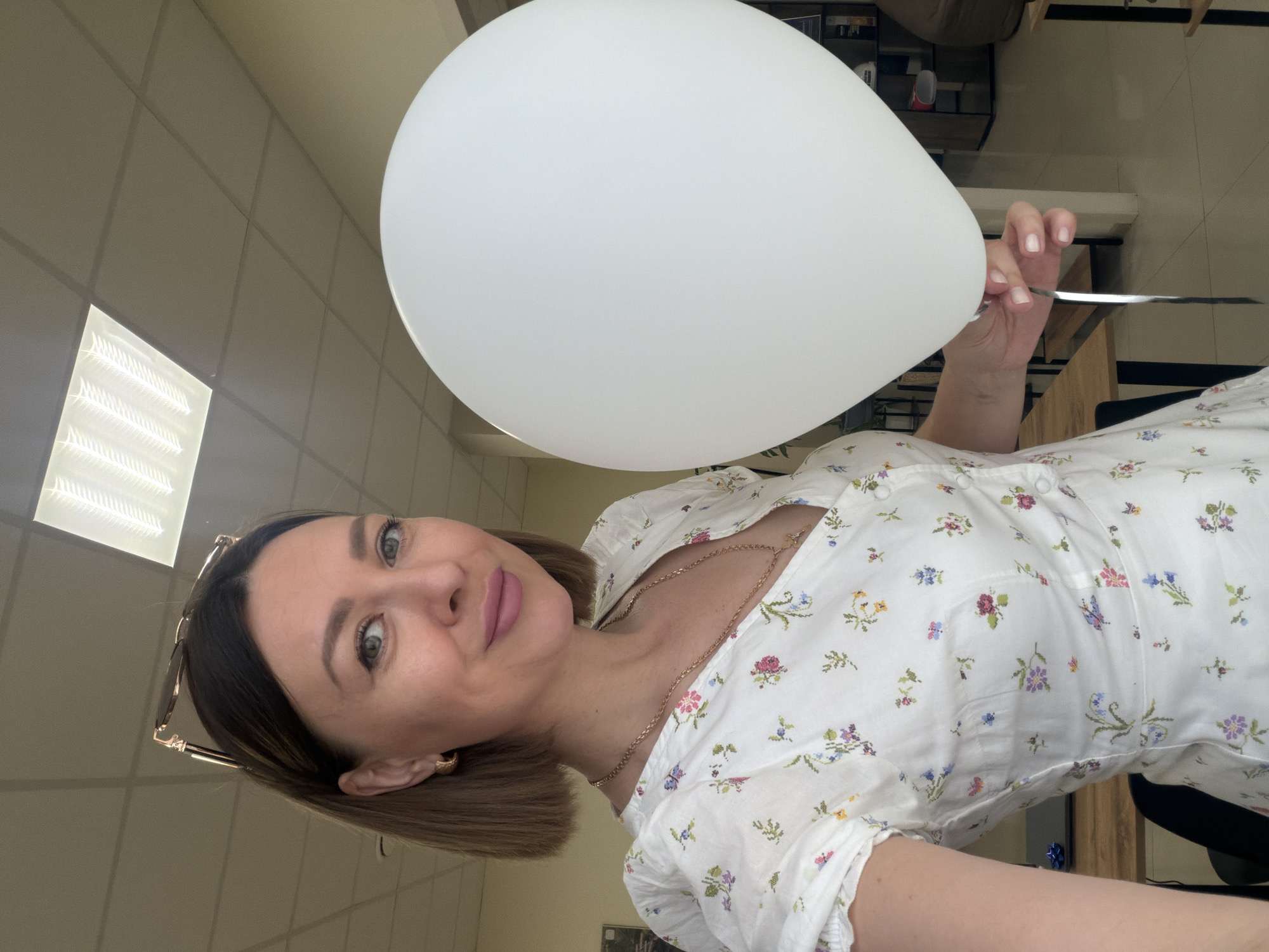 A woman in a white floral dress is holding a large white balloon. She is indoors, possibly in an office, as suggested by the ceiling tiles and fluorescent light fixture. The location in Rostov-on-Don, Russia cannot be confirmed based on the image.