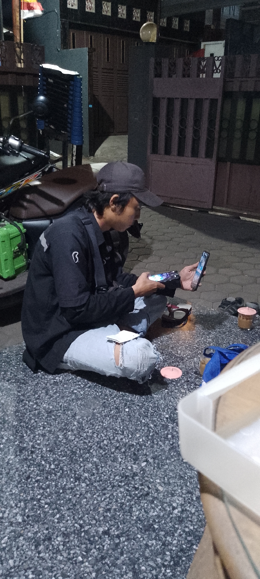 The image depicts a person sitting on a gravel surface outside at night. The person is wearing a black long-sleeved shirt, ripped light-wash jeans, and a dark baseball cap. They are holding two smartphones, looking at the one in their right hand. A motorcycle is partially visible behind and to the left of the person, with a green case attached to it. In the background, there is a closed dark-colored gate with decorative elements and a dark wall with patterned vents. To the right of the gate, there is a darker section of wall and a wooden door. On the ground near the person are a few small items, including what appear to be pink circular objects and a cup with a pink lid containing a brown liquid. A white rectangular object is in the foreground on the right, partially obscuring the view. The overall setting suggests an outdoor area, possibly a residential or communal space in Burikan, Indonesia, given the presence of a motorcycle, residential-style gates, and the paved ground.