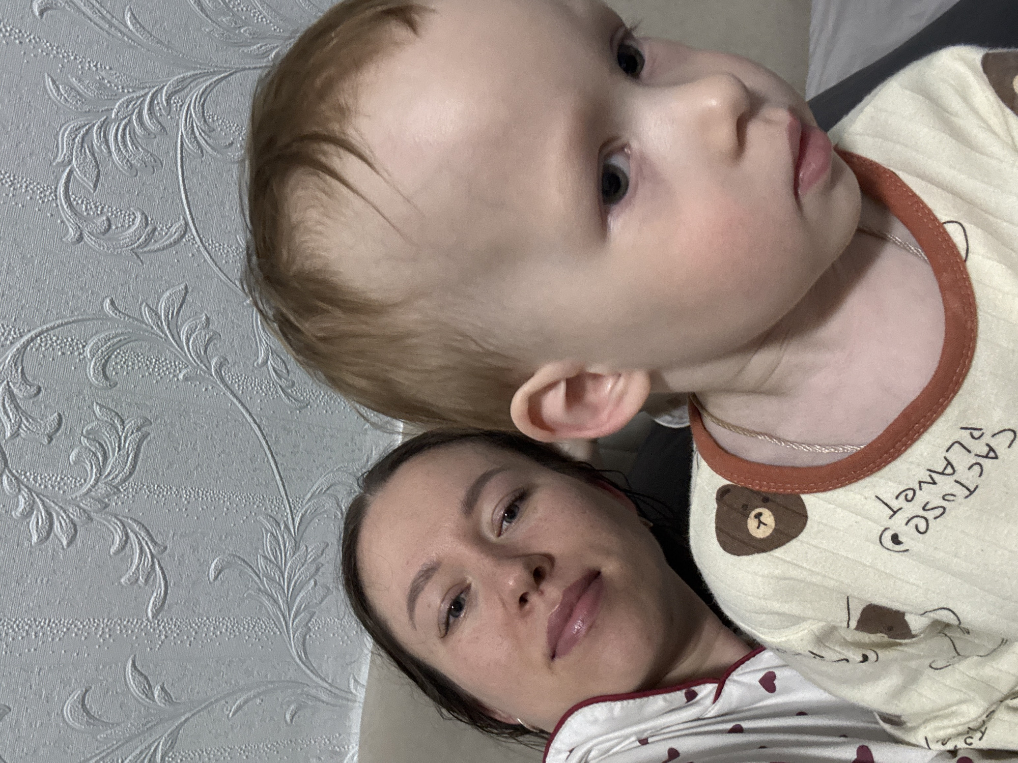 The image shows a close-up view of a woman and a baby, likely indoors. The woman is lying down, and the baby is positioned above her, partially obscuring her face. The woman has fair skin, dark hair, and appears to be wearing light-colored sleepwear with a red trim. The baby has fair skin and light brown hair. The baby is wearing a yellow and brown shirt with an image of a bear and the word "Cactuse Planet". The background consists of a wall with a light-colored patterned design. There is no evidence of the city Ферма or Russia in the scene. The subjects are interacting in a close physical proximity.