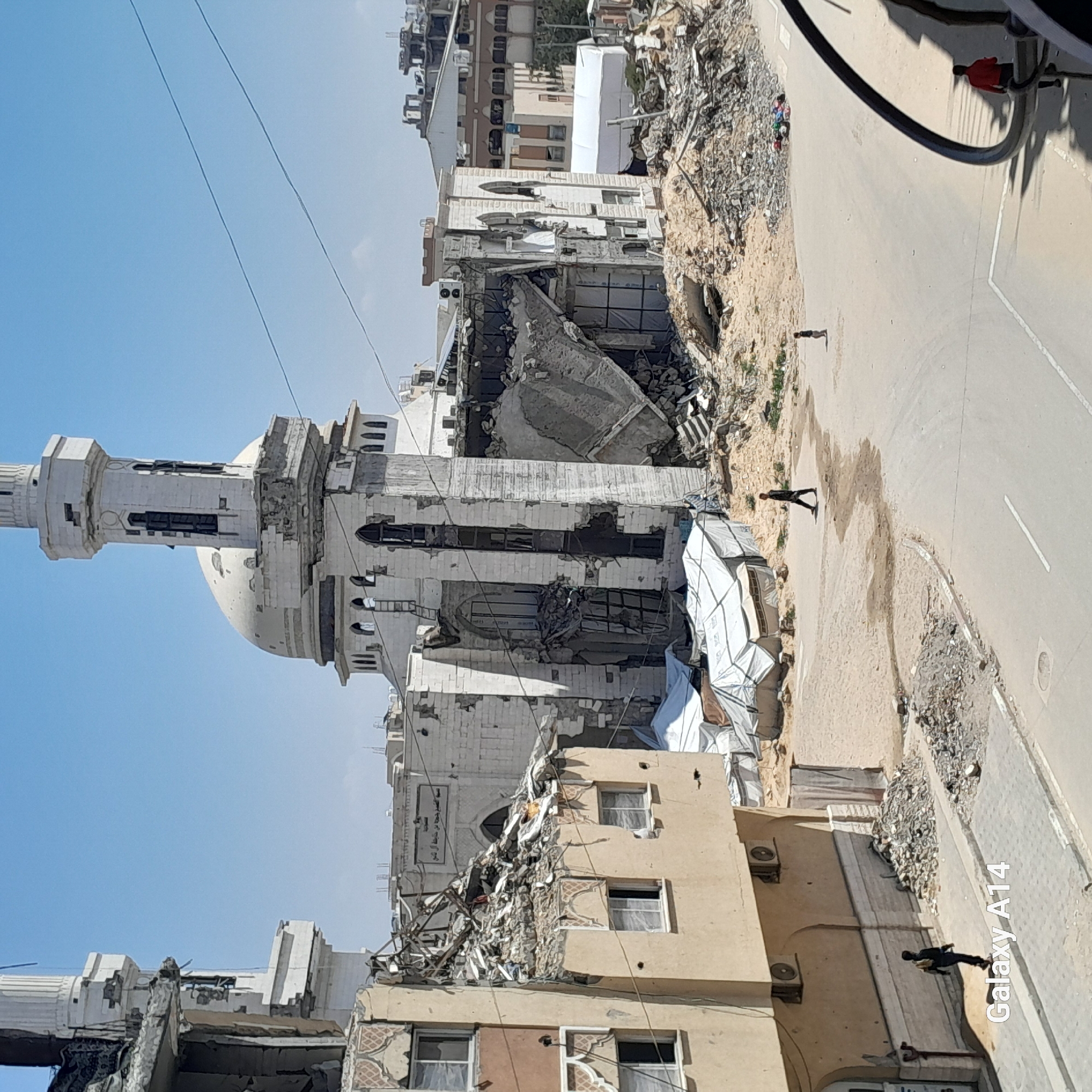 The image shows extensive structural damage to buildings, including a mosque with a prominent minaret and dome, in an urban environment. Debris is scattered throughout the scene. A road runs adjacent to the damaged structures, and people can be seen walking in the vicinity. Tents are set up near the buildings.