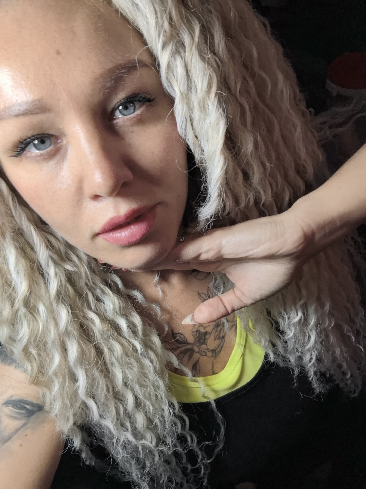 A close-up, high-angle shot shows a woman with light complexion and blue eyes. She has long, curly, blonde hair. Her lips are pink and glossy. She has tattoos on her chest and arm, and is wearing a black and neon green top. Her right hand is placed near her neck. The location is unspecified, and there is no information provided to suggest the photograph was taken in Noviy, Russia.