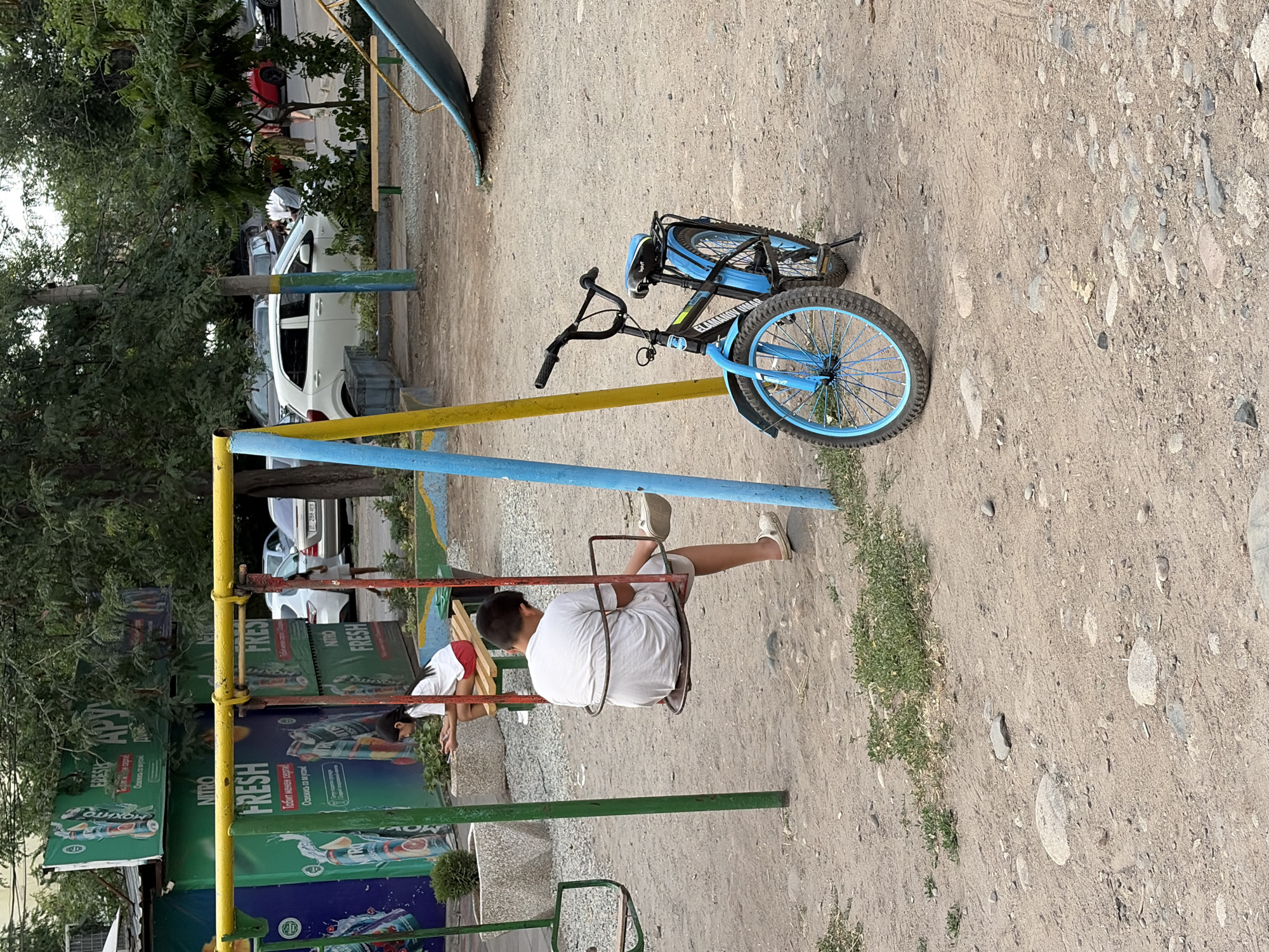 In a sandy playground in an urban area, a young boy, wearing a white t-shirt and shorts, sits on a swing, facing away from the camera. He is wearing white clogs on his feet. To his left, another child, a girl with dark hair, sits on a bench, also facing away, with red shorts and a white t-shirt. A blue bicycle with black handlebars is parked in front of the swing set, leaning on its kickstand. In the background, cars are parked along a street, and trees line the area. Large green banners with advertisements are mounted on structures to the left of the playground.