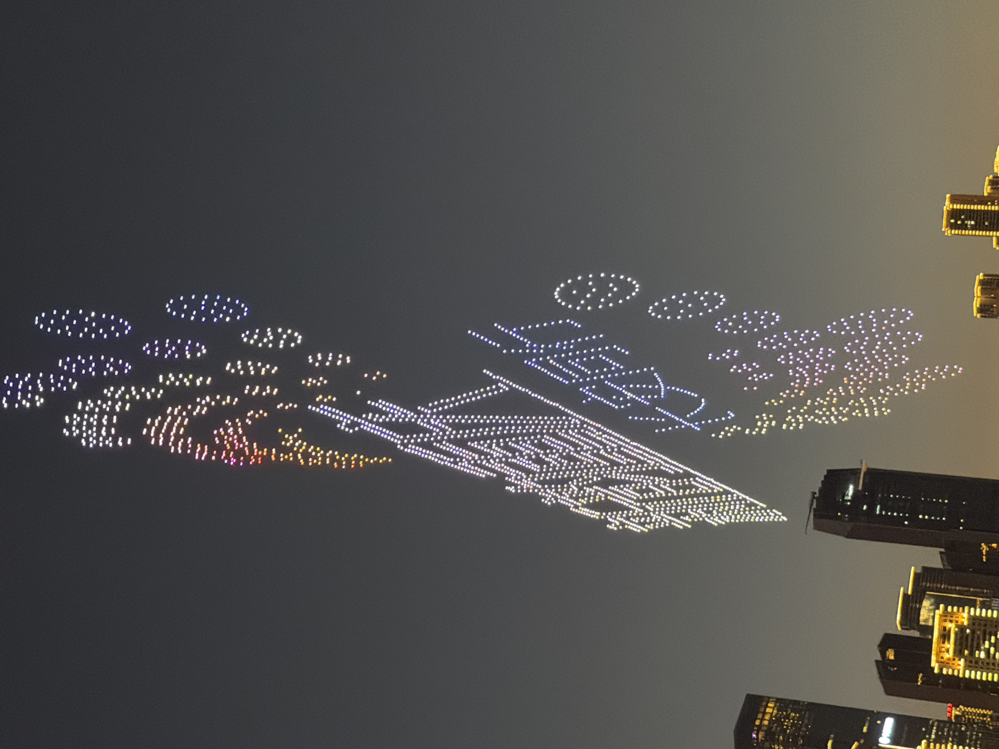The image captures a night-time drone light show above the city of Nan'an District, China. Hundreds of illuminated drones form a large, vertical display in the sky, creating patterns and Chinese characters. The drones are primarily white, with some exhibiting colorful hues of pink, orange, and purple. Below the drone display, the skyline of Nan'an District is visible, featuring several tall buildings with illuminated windows and architectural lighting. The sky is a dark, overcast gray, suggesting dusk or night. The city lights and the drone formations create a vibrant spectacle against the dark backdrop.