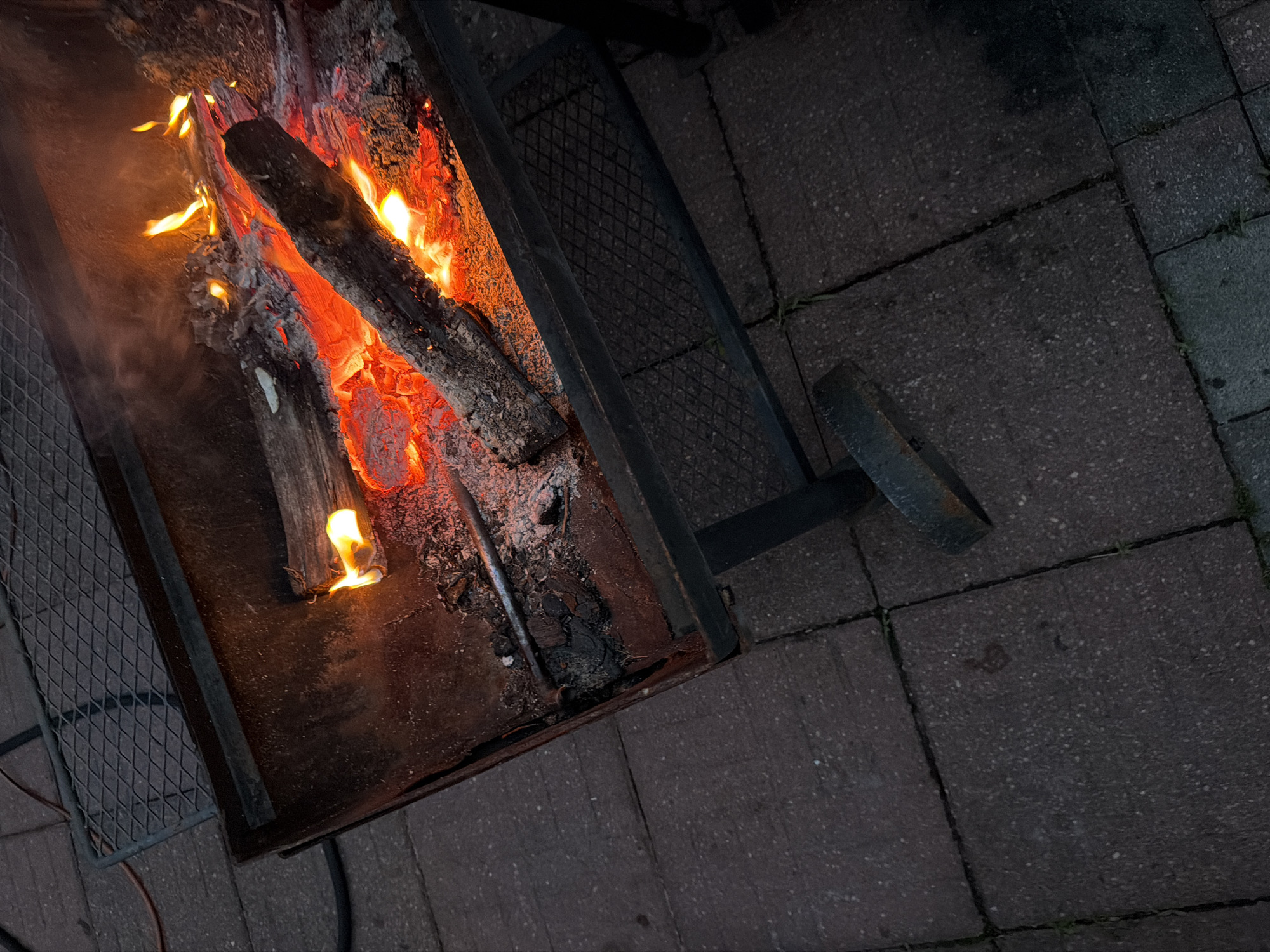 A fire burns in a rectangular metal fire pit, which is supported by a metal frame with legs. The fire pit contains several logs, some of which are actively burning with orange flames and glowing red embers. Ash and soot cover the bottom of the fire pit. The fire pit is situated on a patio paved with dark reddish-brown interlocking bricks. A metal grill rack is visible in the upper left corner of the image, resting on the bricks. The scene appears to be outdoors, and the lighting suggests it is either dusk or nighttime. The location of Riverhead, United States of America, is not discernible from the image.