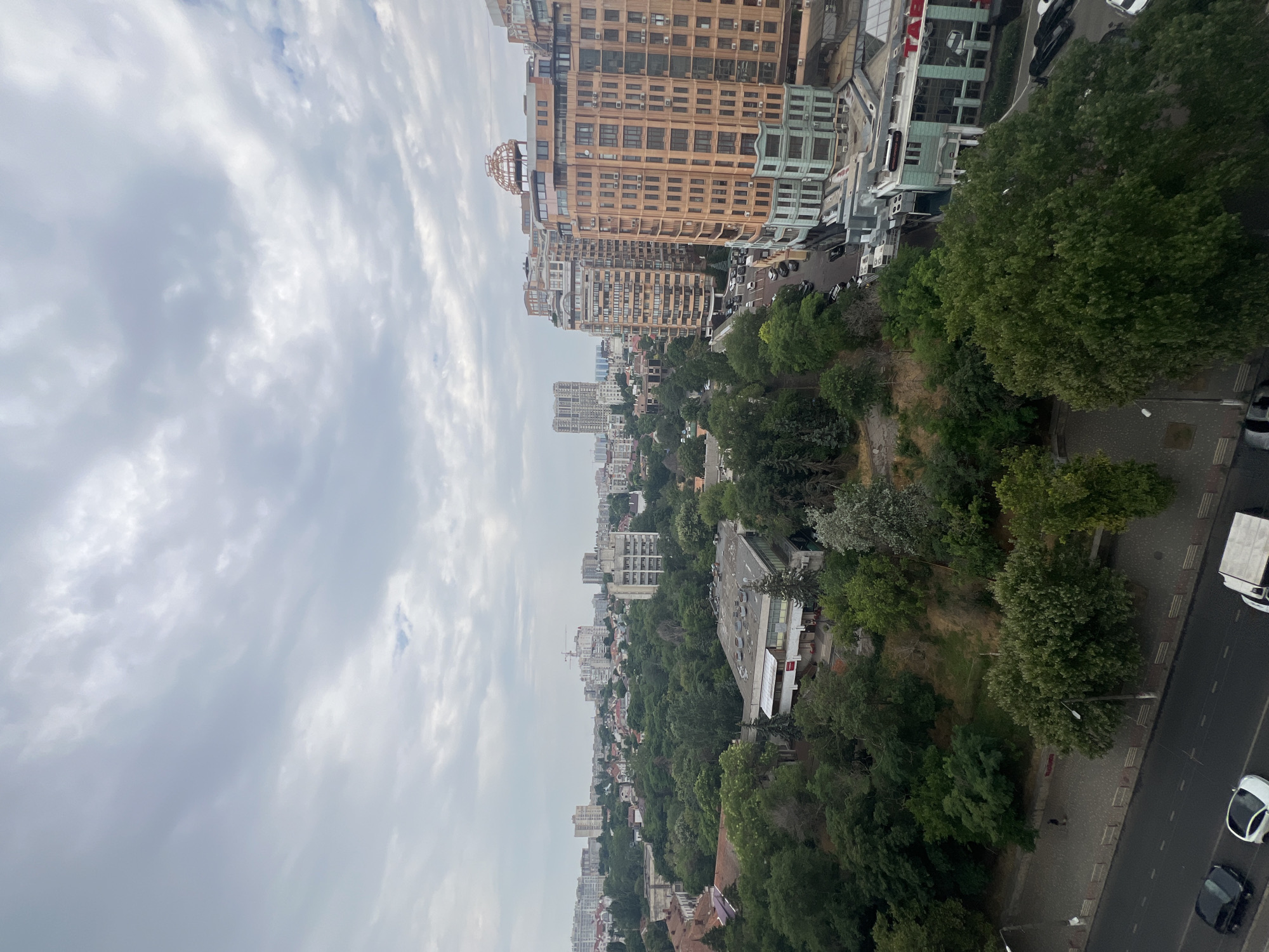 This is a high-angle, exterior, daytime, full shot of a city district in Odesa, Ukraine, with a focus on a street and apartment buildings. The sky is overcast, filled with thick gray clouds. In the foreground, a multi-lane asphalt road is visible, with several cars parked along the side and a few driving. A sidewalk with benches and a street lamp runs parallel to the road. The middle ground is dominated by a dense canopy of lush green trees, interspersed with some lower-rise buildings and a few taller, older apartment blocks. Further in the distance, a cluster of modern, tall apartment buildings stands out, one of which is a distinctive peach-colored building with numerous windows and decorative elements. Construction cranes are visible in the far background, suggesting ongoing development. The overall impression is of a densely populated urban area with significant green spaces.