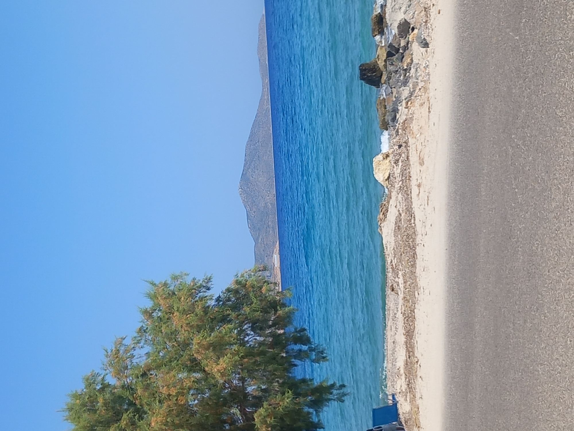 The image depicts a serene coastal scene in Zipari, Greece, under a clear blue sky. In the foreground, a gray asphalt road runs horizontally across the lower part of the frame. Beyond the road lies a sandy beach, scattered with dry seaweed and small rocks. To the left, a single green tree with a dense canopy extends into the frame, its branches partially obscuring the view of the sea. Two blue lounge chairs are visible on the beach, suggesting a place for relaxation. The sea itself is a vibrant turquoise, with gentle waves lapping at the shore. In the distance, a range of low, undulating hills forms the horizon. The overall atmosphere is peaceful and idyllic, characteristic of a Mediterranean summer day.