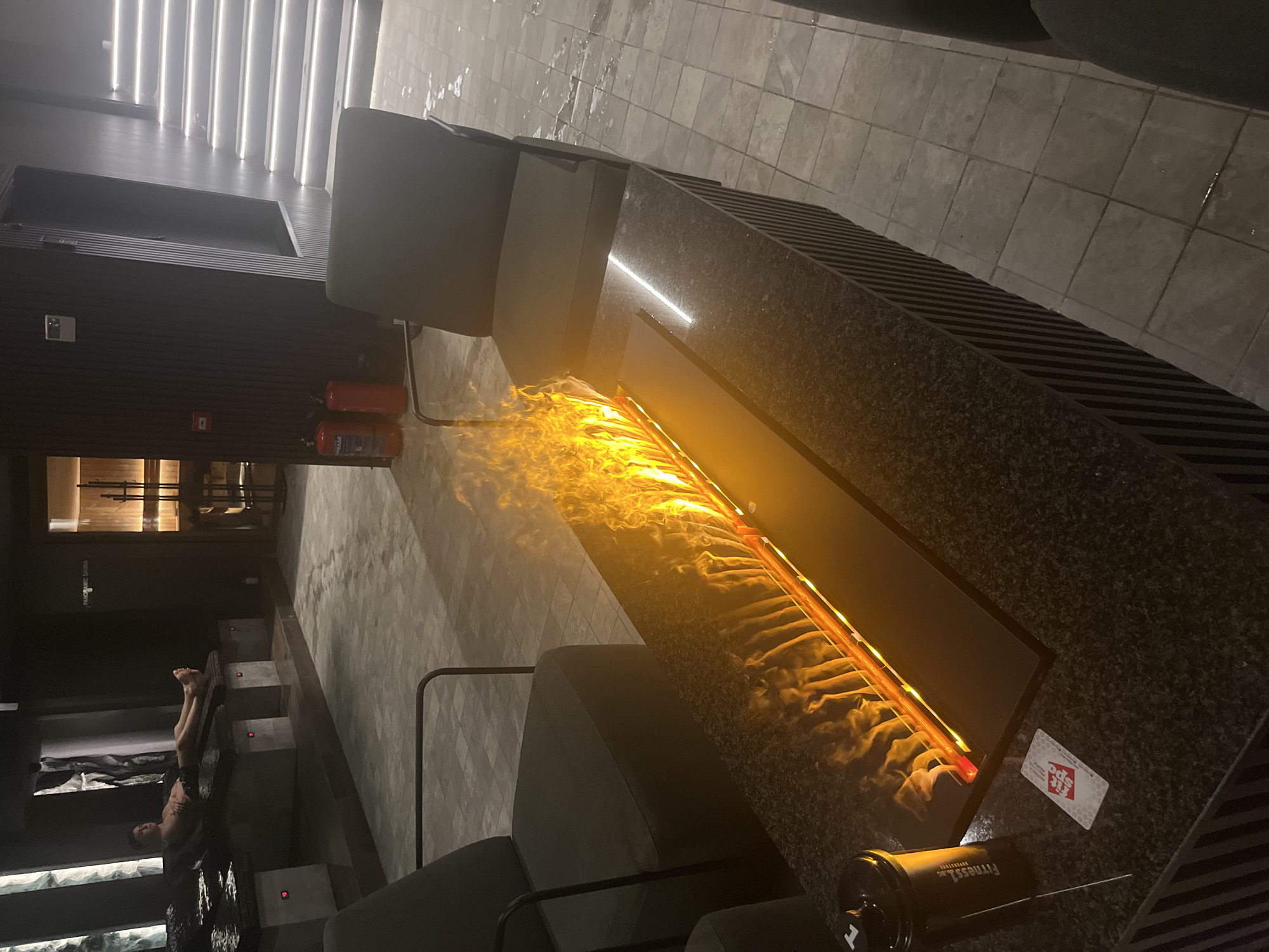 A dimly lit interior shows a panoramic sauna and a fireplace. A person is reclining on steps in the sauna. Two fire extinguishers are on a wall near the sauna entrance. The fireplace is elevated, with flames visible inside a long rectangular opening. Seating is arranged near the fireplace. A "Fit spo" card and a water bottle are on the elevated surface next to the fireplace. The location is likely in Sofia, Bulgaria, due to the name on the card. The floor is tiled. Overhead, there are parallel lights.