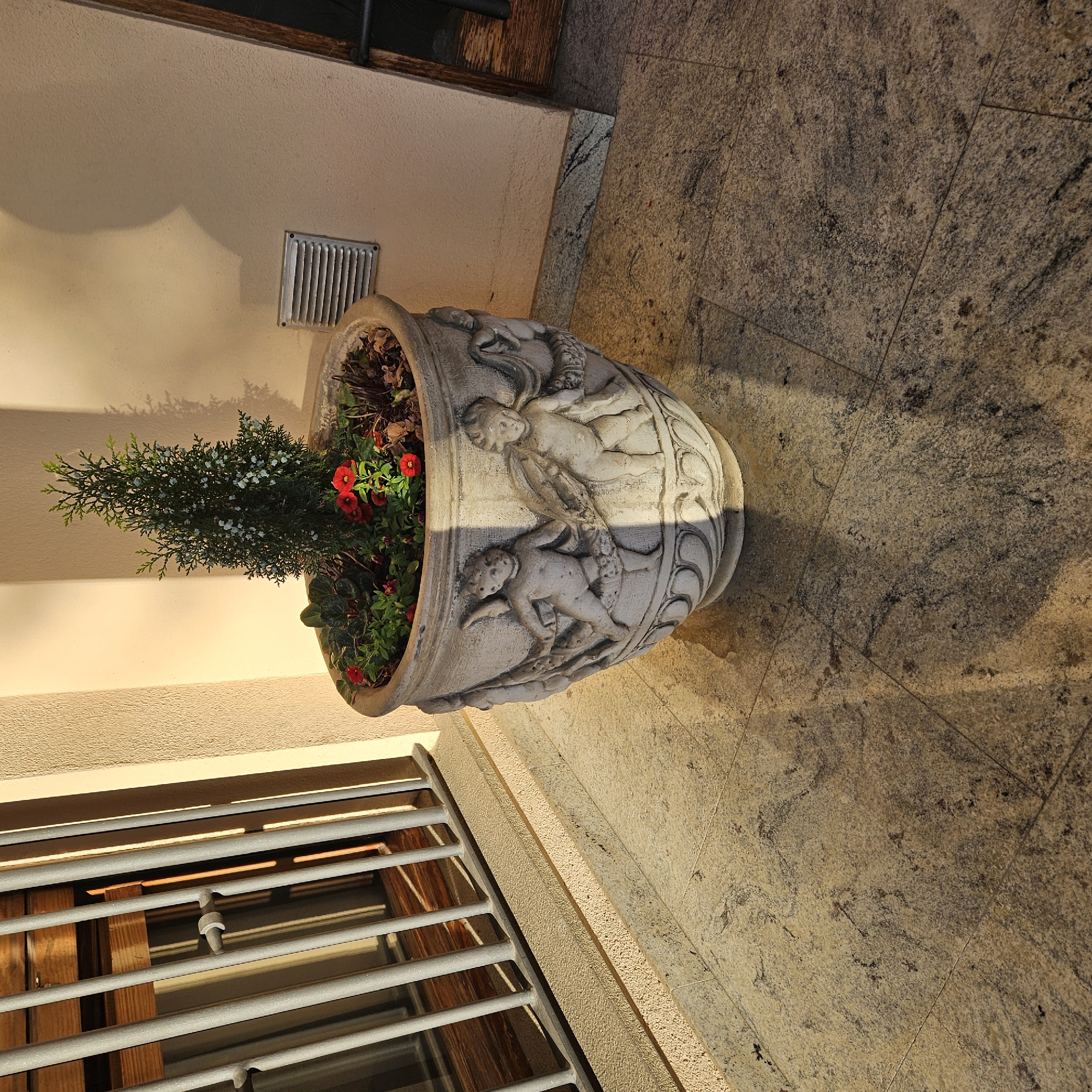 The image shows a decorative stone planter filled with a small evergreen tree and red flowers, placed on a tiled patio. The planter has bas-relief carvings of cherubs on its sides. To the left, a window with metal bars is visible, and to the right, a beige stucco wall with a vent and a wooden door frame are present. The scene is illuminated by natural light, casting shadows. The location is specified as Vilablareix, Spain.