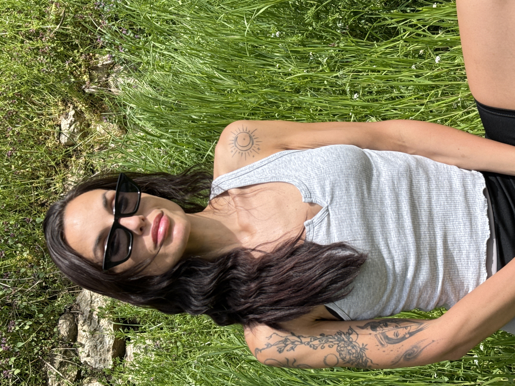 A woman with long, dark, wavy hair is lying in a grassy field. She is wearing sunglasses and a gray tank top, and a black garment on her lower body. Tattoos are visible on both of her arms. The background consists of grass, vegetation and some stones. This image was taken outside of an urban area, but a precise location in Pleven, Bulgaria is indiscernible.