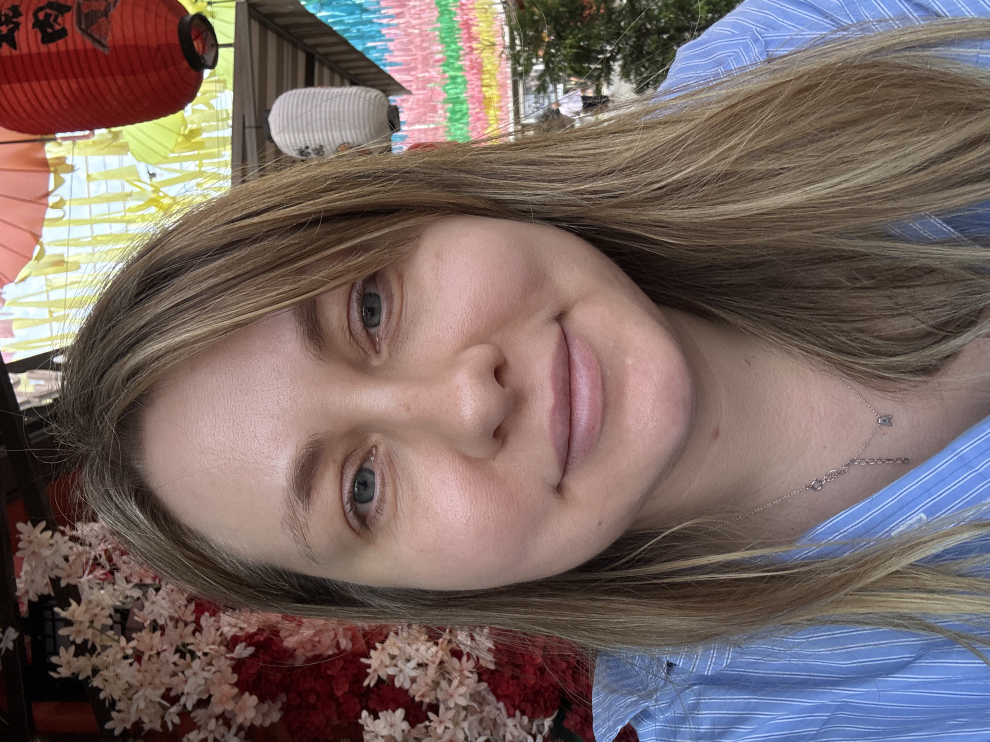 A Caucasian woman with blonde hair and blue eyes is pictured in a selfie. She is wearing a blue and white striped collared shirt and a silver necklace with a small pendant. The background appears to be outdoors, with elements that suggest an Asian cultural setting, including red and white lanterns and colorful banners. There are also floral decorations, possibly cherry blossoms, visible. The woman is looking directly at the camera and smiling slightly. No specific location in Krasnodar, Russia, can be identified from the image.