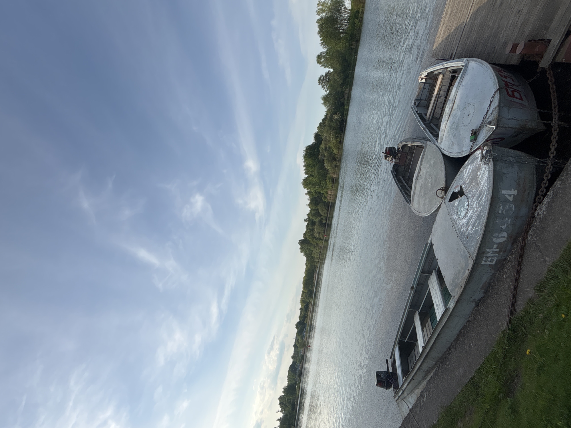 The image shows several small boats docked along a concrete bank of the Svislach River, likely in Minsk, Belarus. The boats are made of metal and appear to be of a similar design. They are painted in shades of gray and silver and show signs of wear and use. Some of the boats have outboard motors attached. The water is calm and reflects the sky, which is partly cloudy. Trees line the opposite bank of the river. A wooden dock is visible near the boats. The overall scene suggests a quiet, natural setting within an urban environment.