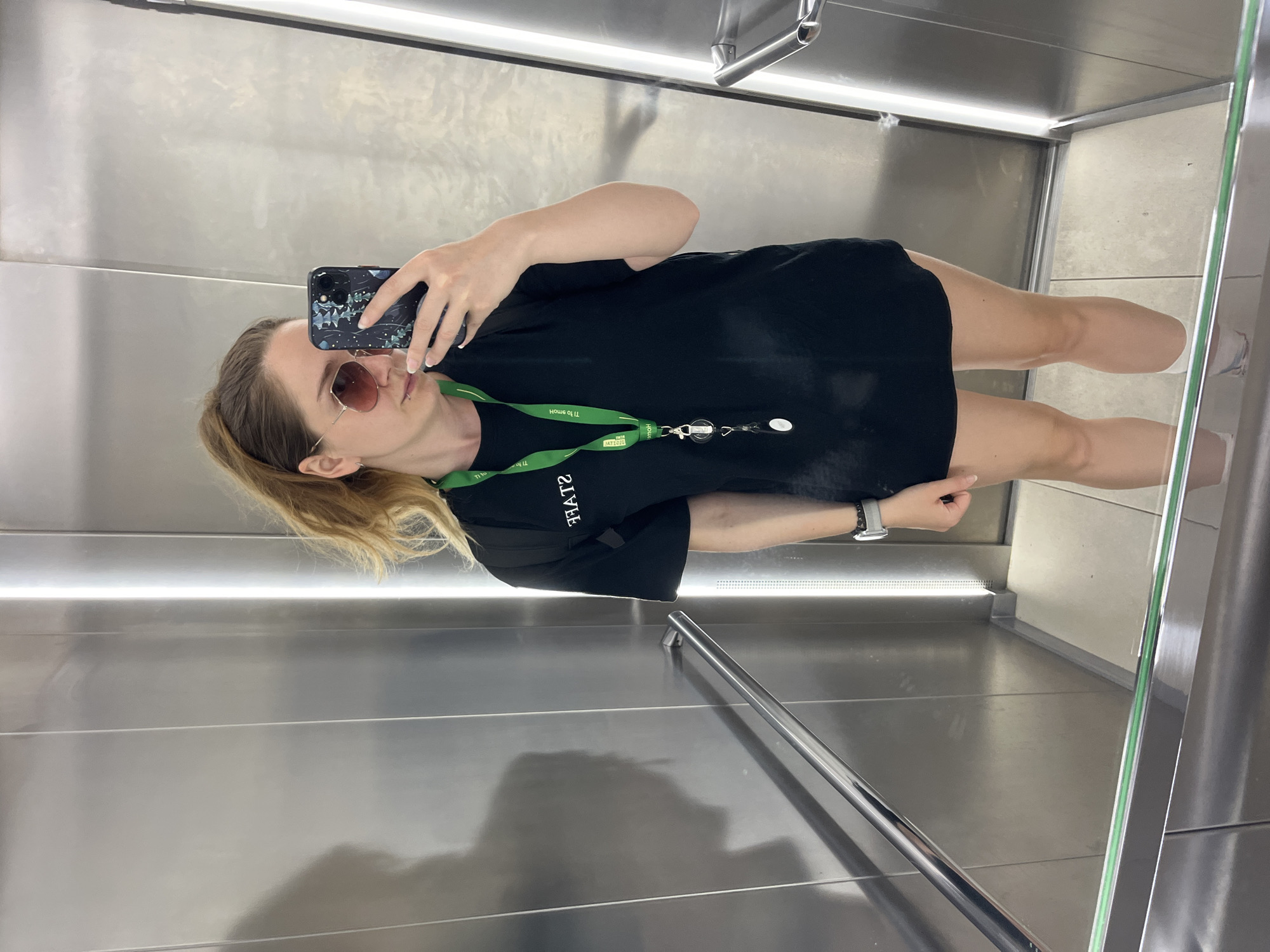 A woman is taking a mirror selfie in an elevator. She is wearing a black t-shirt with "STAFF" printed on the back, sunglasses, and a lanyard with an ID card. The elevator walls are made of polished metal, and there is a mirror at the bottom. The location is not specified in the image or metadata, therefore it cannot be determined if it is in Plovdiv, Bulgaria.