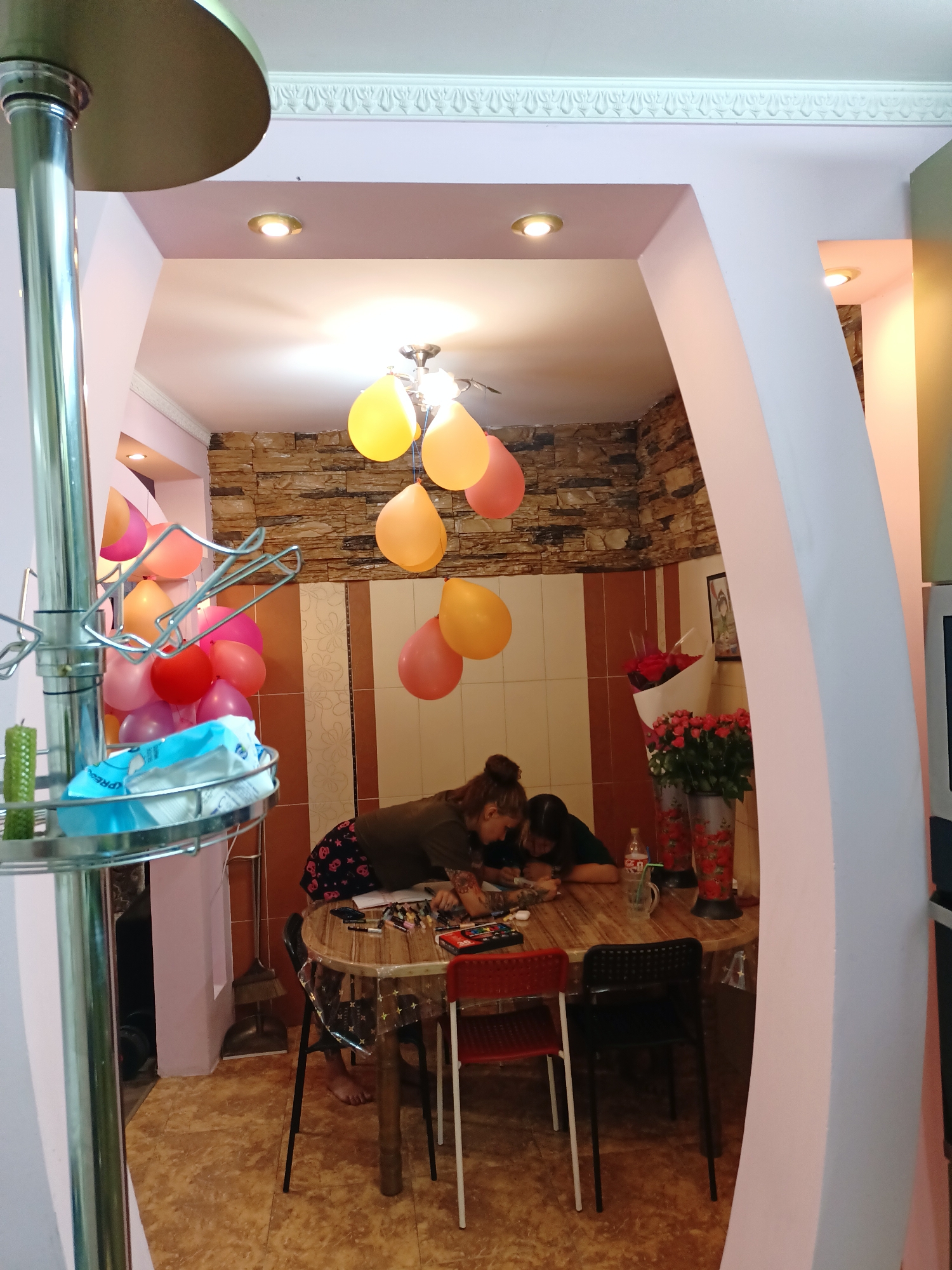 The image depicts an interior scene, likely a domestic setting, with a strong emphasis on decorations. A prominent feature is the presence of numerous colorful balloons, primarily yellow, orange, and pink, suspended from the ceiling. The walls appear to be decorated with stone and patterned tiles. Two individuals, both appearing to be female, are seated at a round wooden table. One is wearing a dark, patterned garment, while the other is in a dark-colored top. They are both leaning over the table, engaged in an activity that involves looking at items on the table, possibly art supplies or papers. Several chairs surround the table, some of which are red and black. In the background, a bouquet of red flowers in vases is visible. To the left of the frame, a metal stand holds a green cylindrical object and a blue and white packaged item. The lighting in the room is warm, with recessed ceiling lights illuminating the space. The floor is tiled with a mottled brown pattern. The overall impression is that of a celebratory or decorated interior space.