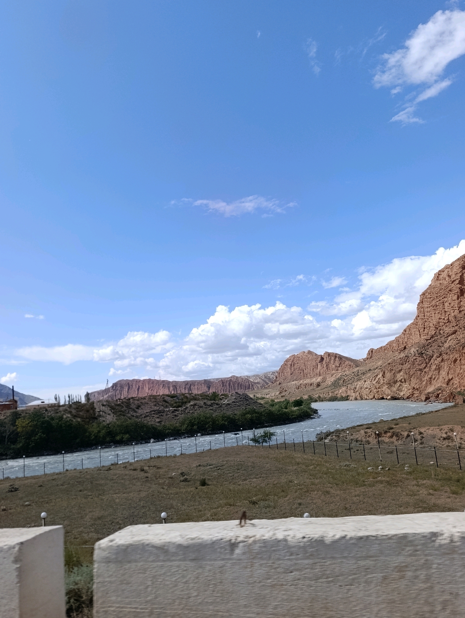 The image captures a scenic view of the Naryn River in Naryn City, Kyrgyzstan. The river flows through a valley flanked by rugged, reddish-brown mountains. The sky is a clear blue with scattered white clouds. In the foreground, there is a dry, grassy embankment with a fence and some lampposts. In the far left background, there appears to be an industrial building or factory. The overall scene depicts a natural landscape with human infrastructure present.