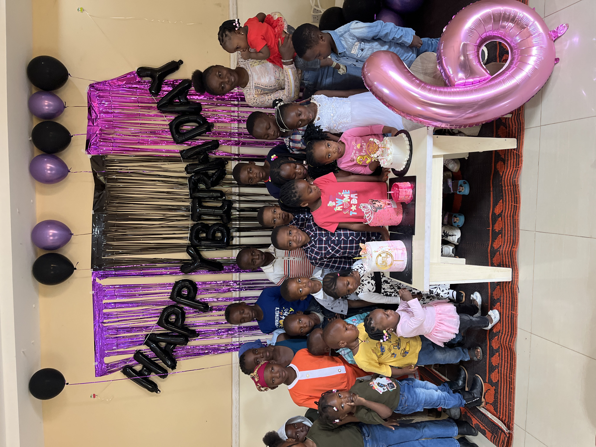 The image depicts an indoor birthday celebration in Kuru Karama, Nigeria. Approximately 20-25 individuals, predominantly children ranging from toddlers to young teenagers, are gathered. An adult female, positioned on the right, holds an infant clad in a red outfit. The group is arranged around a light-colored rectangular table displaying three birthday cakes and a smaller, dark pink cylindrical item, possibly another small cake or candle holder.
The cakes vary in design: the leftmost is pink, possibly multi-tiered, featuring a cartoon character (possibly a princess) and a crown. The middle cake is smaller, light pink with a single burning candle. The rightmost cake is white with pink frosting accents, topped with a gold "Happy Birthday" inscription and a butterfly decoration. A small child in a pink tutu dress, positioned in front of the table, extends an arm towards the cakes, indicating engagement.
The background features a cream-colored wall adorned with purple and black metallic fringe curtains. Black foil balloons spelling "HAPPY BIRTHDAY" are affixed above the curtains. Several black and purple latex balloons float near the ceiling. On the tiled floor, to the lower right, a large pink foil balloon shaped like the number "6" rests. The floor is partially covered by a dark patterned rug with orange and black geometric designs. Most individuals are looking directly at the camera, some smiling, contributing to a celebratory atmosphere. The attire is casual and colorful, with children wearing t-shirts, jeans, and dresses.