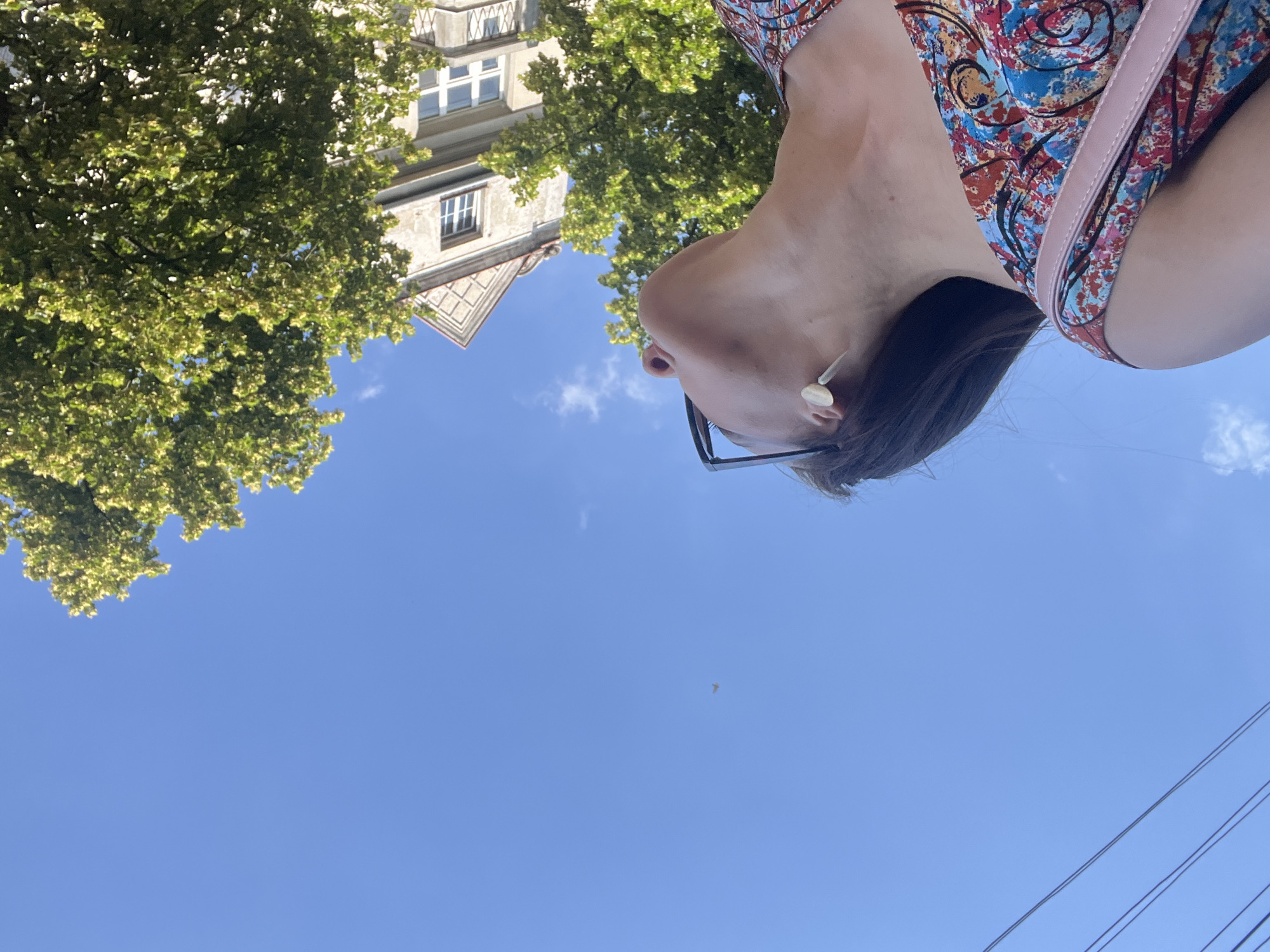The image is an outdoor, medium shot of a woman viewed from a low angle, looking up towards the sky. She is wearing a colorful, patterned dress with shades of red, blue, and brown. A pink strap from a bag is visible over her shoulder. She has dark hair pulled back, and is wearing earrings and eyeglasses. The background features a bright blue sky with scattered white clouds. To the upper left of the frame, there is a building with white-trimmed windows and a tree with vibrant green foliage. The lighting suggests it is daytime, likely sunny. The woman's gaze is directed upwards, and her expression is neutral.
