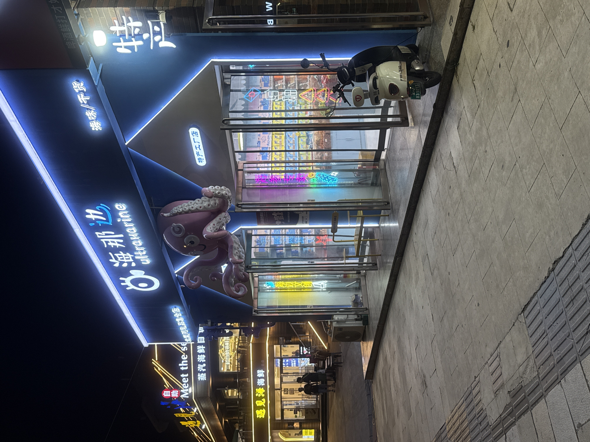The image shows a storefront at night in Qinhuangdao, China, identified by the text "遇见海鲜" (Meet the Sea) and other Chinese characters. The storefront features prominent neon blue signage with the words "海那达 ultramarine" and an illuminated octopus decoration above the entrance. To the right of the entrance, a white and black scooter is parked on the sidewalk. The sidewalk is paved with light-colored tiles and features a tactile paving strip. In the background, other illuminated storefronts are visible. The scene appears to be a commercial street or market area.