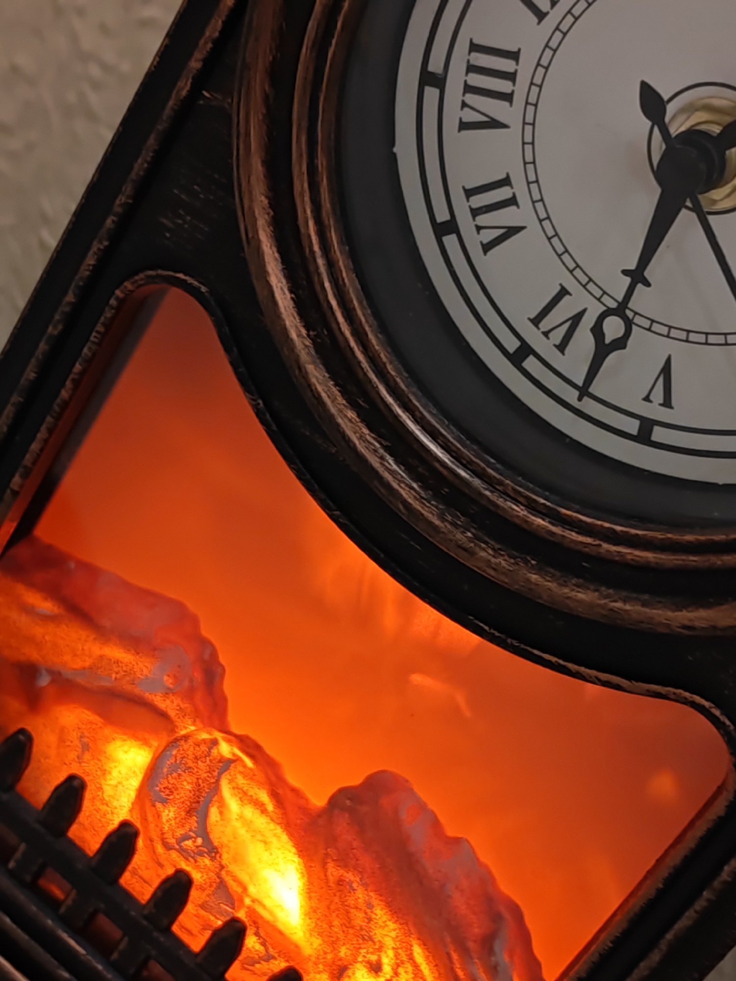 The image shows a close-up of a decorative clock with a faux fireplace beneath it. The clock face is white with Roman numerals, and its hands point to approximately 7:52. The faux fireplace contains illuminated faux logs that glow with an orange and yellow light, mimicking flames. The clock has a dark, textured frame with bronze accents. The background is out of focus and appears to be a textured wall. There are no people or animals present in the image, and no specific location in Bataysk, Russia can be identified from the visual information provided.