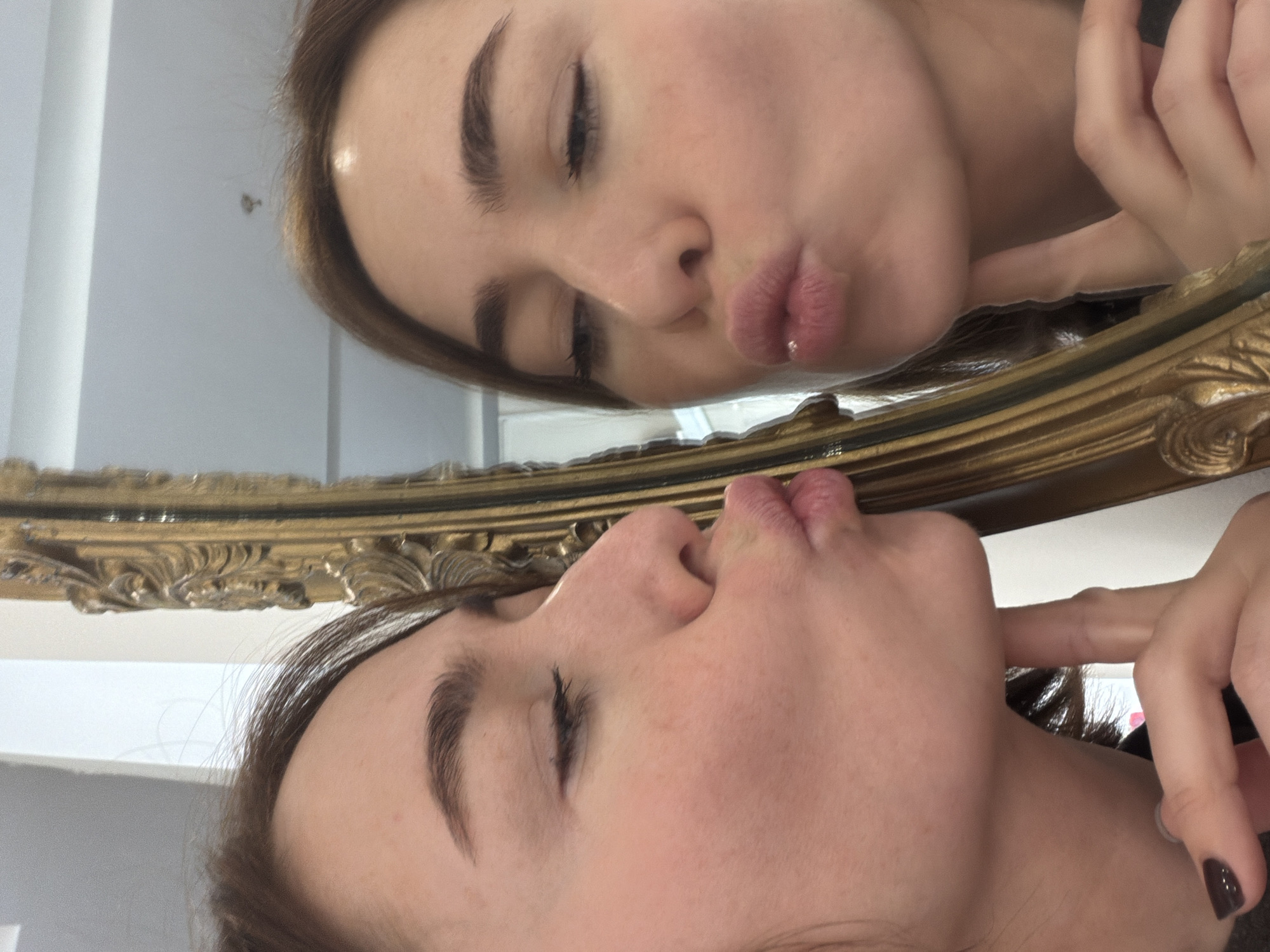 A selfie-style, close-up image depicts a person with brown hair and fair skin, reflected in a gold-framed mirror. The person is making a kissing face. The background includes a white wall. No information is available to confirm the location of this image.