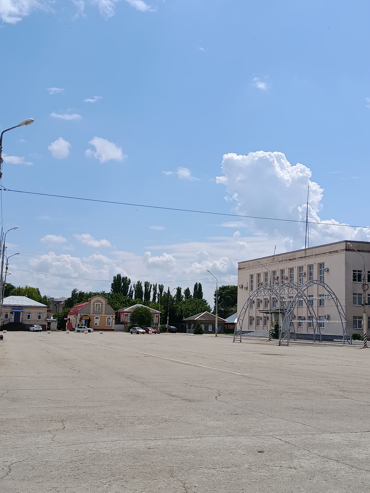The image depicts a street scene in the city of Marx, Russia. In the foreground is a large, paved area, possibly a public square or parking lot, with visible cracks and lines in the concrete. Beyond this area, several buildings are visible. On the left, a light-colored building with a dark-tinted entrance is situated, with a parked white car in front. Next to it is a narrower, light-colored building. Further to the right, a two-story building with a red roof and yellow facade stands prominently. To the right of this, another building with a red roof is visible, with a dark-colored car parked in front and a red car behind it. In the background, a row of tall, dark green trees forms a natural border. To the far right, a larger, light-beige building with numerous windows and air conditioning units is prominent. In front of this building, a modern, metallic arched structure is erected. The sky above is a bright blue with scattered white cumulus clouds. A lamppost is visible on the left side of the image. The scene appears to be captured on a sunny day, likely in the daytime. No people are clearly visible in the image, and there are several cars parked in various locations.