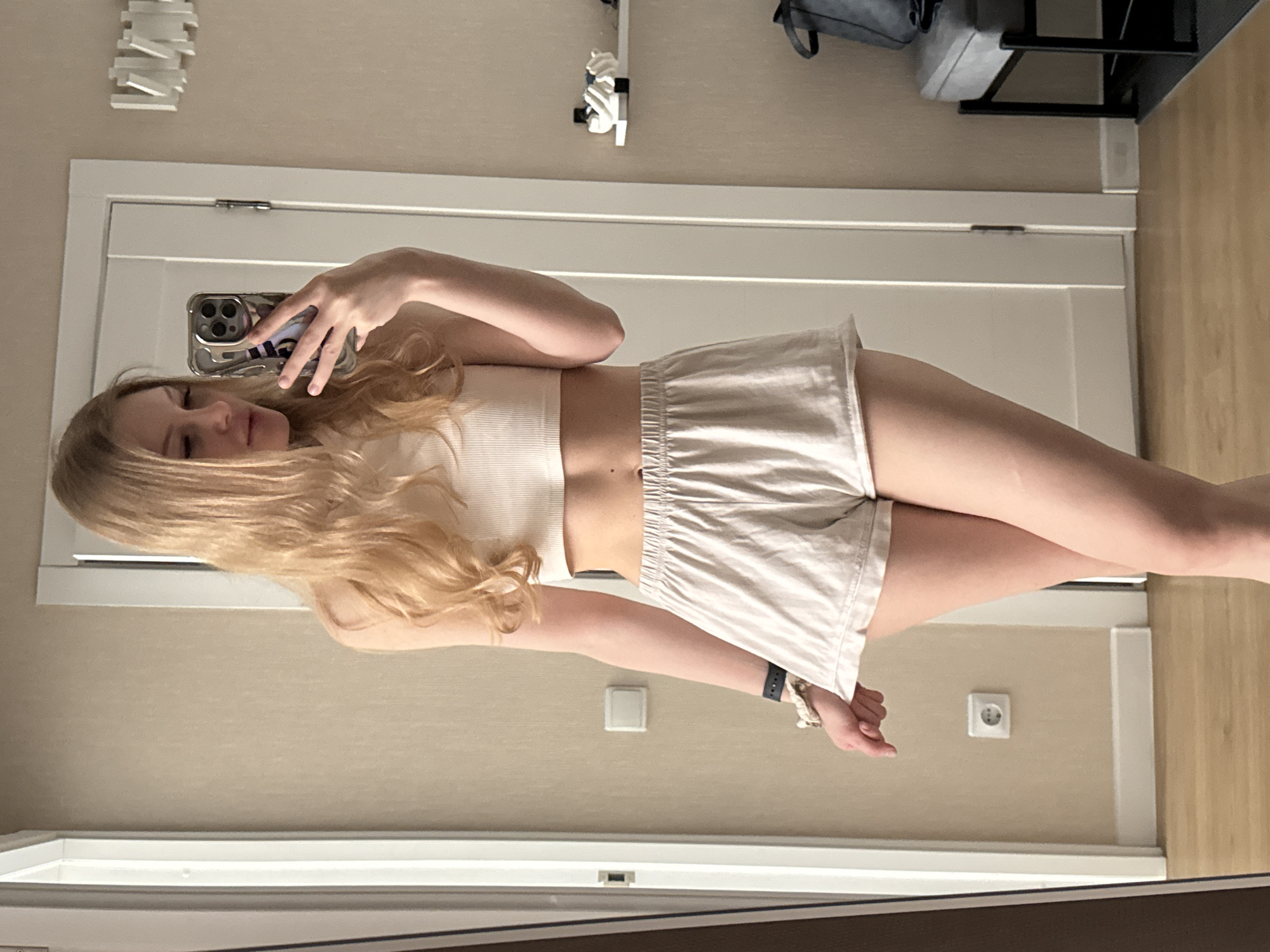 A woman with blonde hair takes a selfie in front of a white door. She wears a white ribbed crop top and beige shorts. The background includes an off-white wall and a glimpse of flooring, possibly in an apartment or home setting. A decorative item and a rack are on the wall behind her, in an area above the door. The electrical outlet suggests this photo was taken indoors. The image was likely captured somewhere within Russia, although it is not possible to determine the precise location without further information.