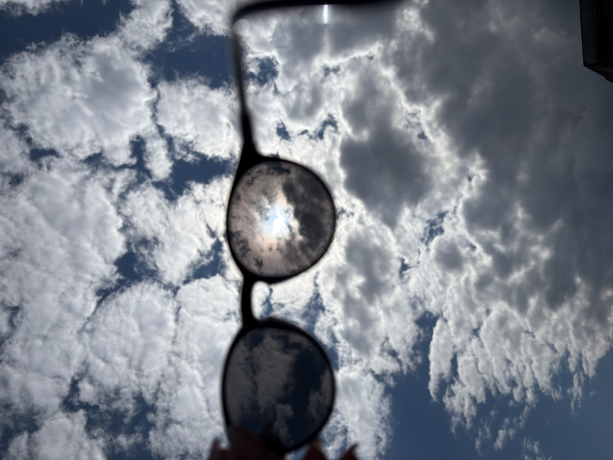 An upward-facing, low-angle shot captures a pair of sunglasses held against a cloudy sky, likely in Chaikovsky, Russia. The central focus is the dark-framed sunglasses, with their lenses reflecting and filtering the sky and sun.
Through the upper lens, the sun appears as a bright, white-blue hotspot, partially obscured by clouds, creating a striking glare. The lower lens reflects a darker, more muted view of the clouds. The silhouette of the sunglass frame and possibly a strap extends upwards, while at the very bottom, parts of fingers can be seen holding the glasses.
The sky itself is a mix of bright white, fluffy cumulus clouds and darker, denser grey clouds, interspersed with patches of clear blue sky. The lighting suggests it is daytime, likely bright daylight given the intensity of the sun visible through the lens. There is no discernible activity beyond holding the sunglasses up to the sky. No visible text or specific location cues beyond the provided context are present in the image.