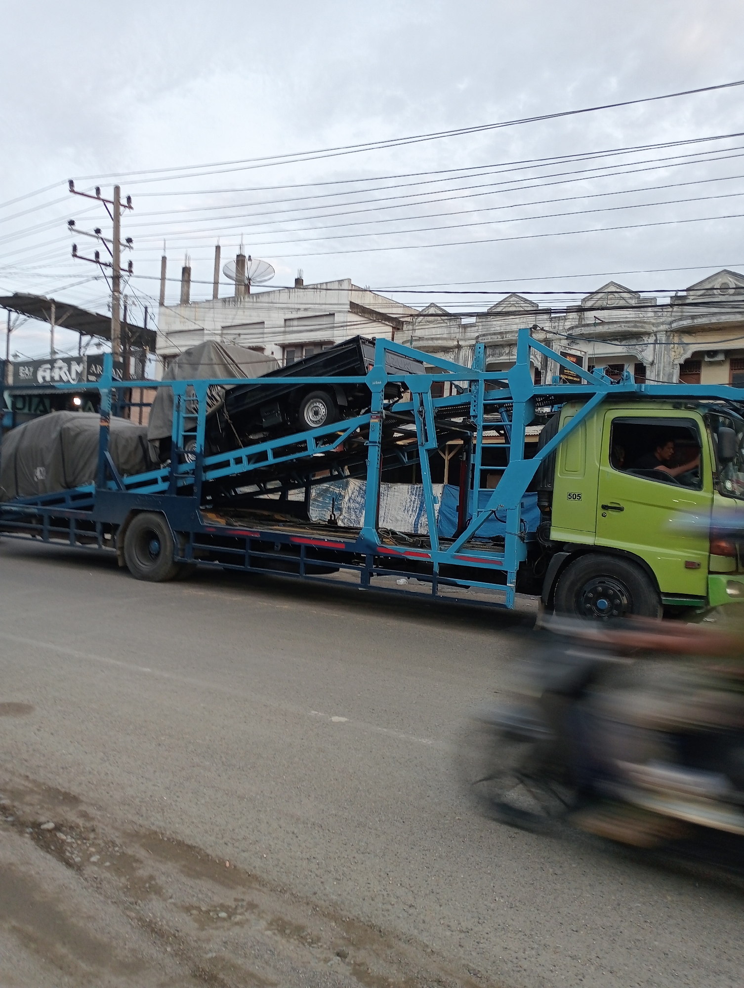 A car carrier truck is moving on a road in Tingkeummanyang, Indonesia. The truck, with a lime green cab and blue framework, carries a black vehicle on its upper level and covered items on the lower level. A blurred motorcycle passes in the foreground. In the background, utility poles and buildings are visible under a cloudy sky.