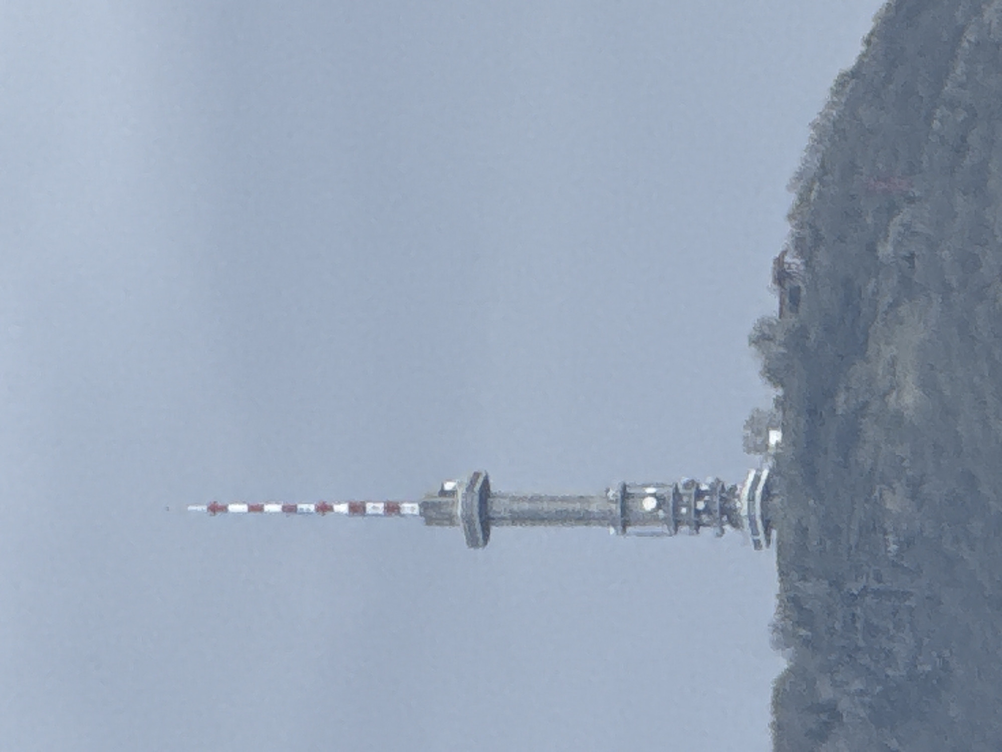 The image depicts the Kopitoto TV Tower in Sofia, Bulgaria, situated atop Mount Vitosha. The tower is prominently visible against a backdrop of a cloudy sky and the mountainside. The tower itself has a cylindrical structure with a distinctive red and white striped antenna. It is located next to a mountain.