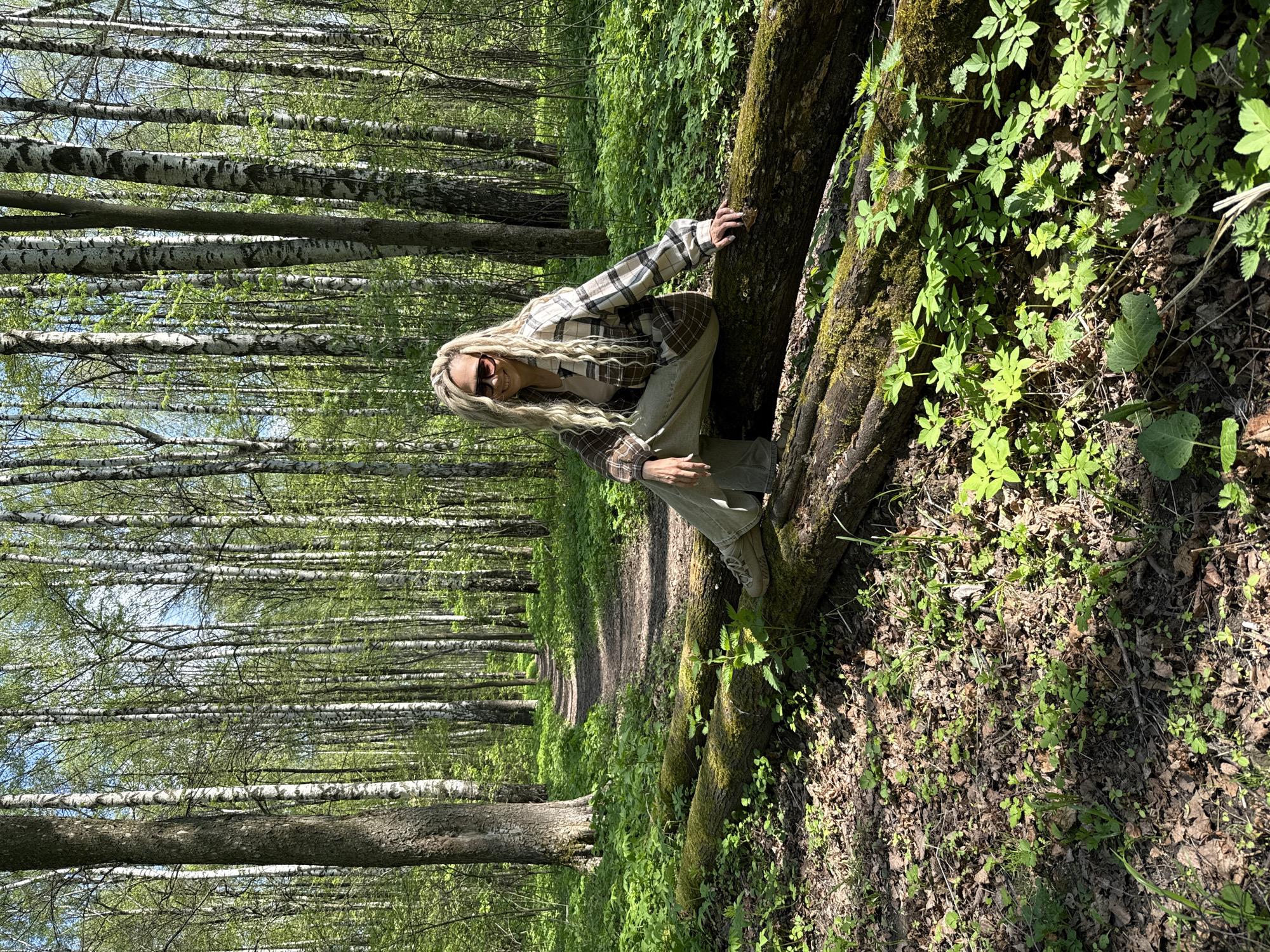 The scene shows a forest with numerous birch trees, identified by their white bark and green leaves. A woman is positioned atop a moss-covered fallen tree. She wears sunglasses, a plaid shirt over a light-colored top, and trousers, squatting on the tree trunk. A dirt path is visible, extending into the depth of the forest behind her. The ground is covered with fallen leaves and greenery, indicative of a wooded area. The location is Rusanovo, Russia.