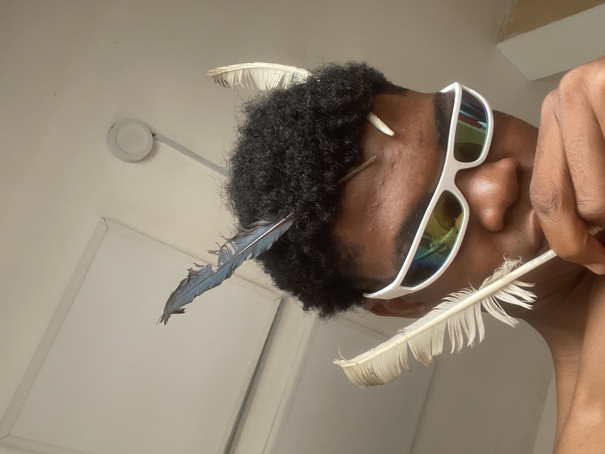 The image shows a close-up of a Black person's head and shoulders, with a focus on their face and hair. They are wearing white, futuristic-style sunglasses with mirrored lenses that reflect a colorful scene. Several bird feathers are placed in their curly, dark hair, with one large white feather prominently displayed near their cheek, held by their hand. Another blue-tinged feather is visible on the top of their head. The background appears to be an indoor environment with a light-colored ceiling that has a visible fixture and a corner of a white panel. There is also a hint of a wall with some texture or discoloration on the right side of the frame. The lighting suggests an indoor setting, possibly with natural light coming from an unseen source. The person's skin tone is medium to dark brown. The image is taken from a low angle, looking up at the subject. The scene does not provide any specific indicators of a location within the city of Ado, Nigeria.